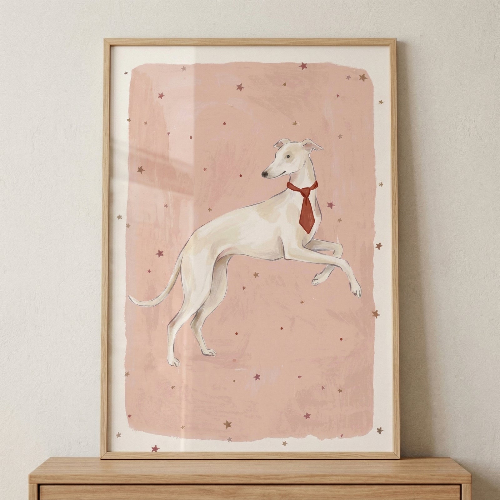Framed illustration of a slender white dog mid leap wearing a red necktie against a soft pink background with scattered stars. The playful pose and simple pastel style create a charming and whimsical nursery wall art piece.