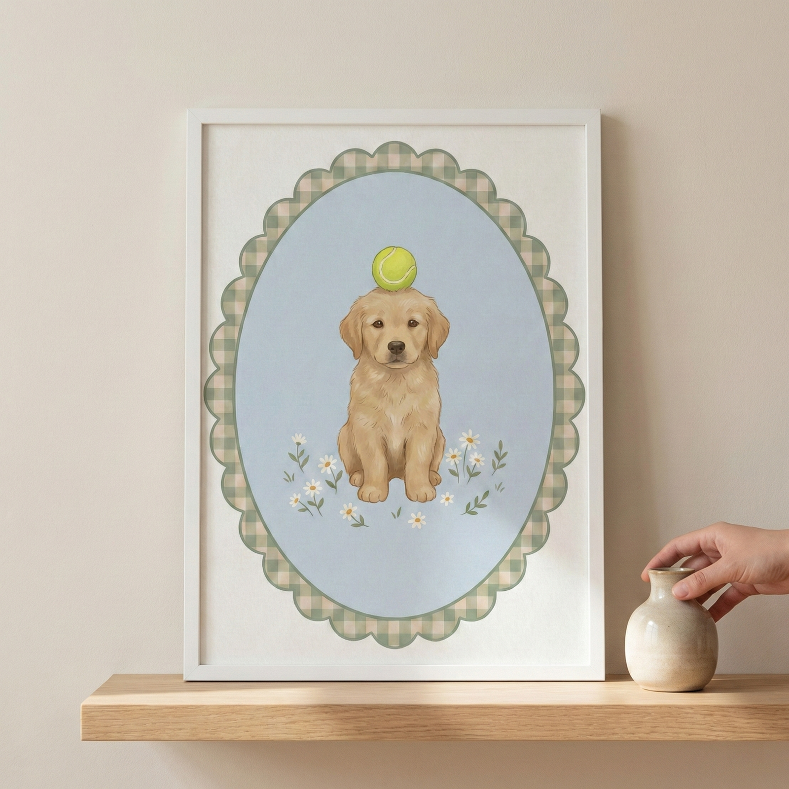 An illustration of a light brown puppy sitting patiently with a green tennis ball balanced on its head. The puppy is set against a light blue oval backdrop decorated with small white flowers. Surrounding the oval is a scalloped border with a green and white checkered pattern. The entire artwork is presented in a plain white frame, which sits on a wooden shelf next to a small, rounded ceramic vase.