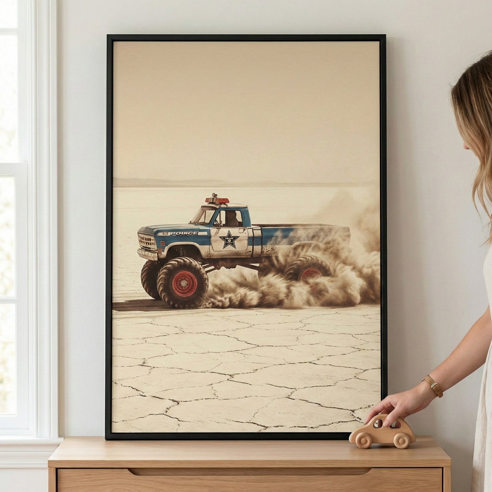 A framed vertical poster of a vintage-style blue and white pickup truck with oversized monster tires, styled as a law enforcement vehicle with a star emblem and a roof-mounted light bar. The vehicle is depicted driving across a vast, cracked desert landscape, kicking up a large cloud of dust. The artwork is hanging on a plain wall above a wooden dresser, where a person’s hand is visible resting near a small wooden toy car.