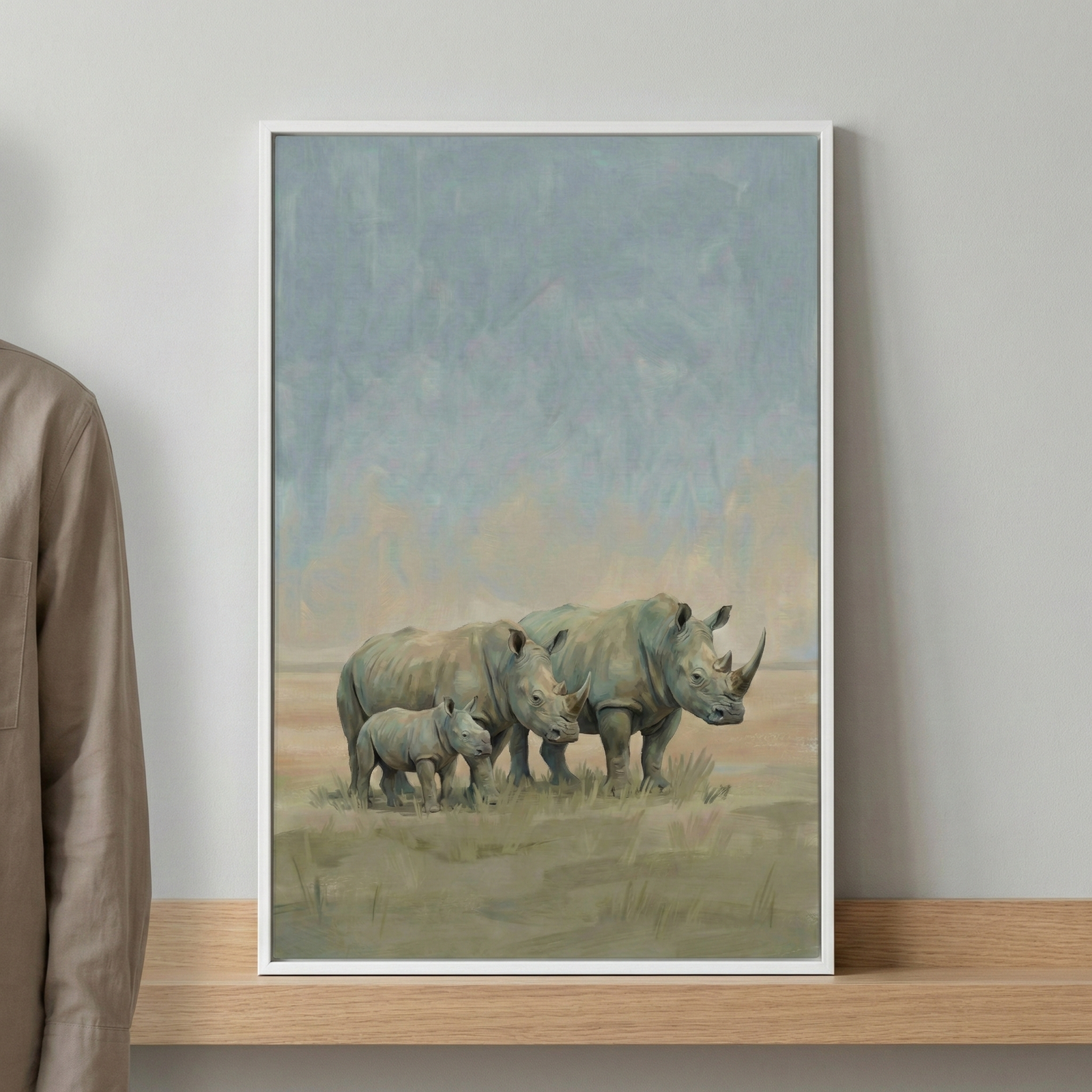 A vertical art piece in a white frame sits on a wooden shelf against a neutral grey wall. The artwork depicts a group of three rhinoceroses—two adults and a small calf—standing together in a grassy landscape. The sky above them is rendered in soft, blended tones of blue, grey, and hints of pale yellow, giving a sense of vastness. The animals are depicted with realistic textures and muted grey and brown tones, suggesting a tranquil scene in nature. The overall style of the art features visible brushwork and 