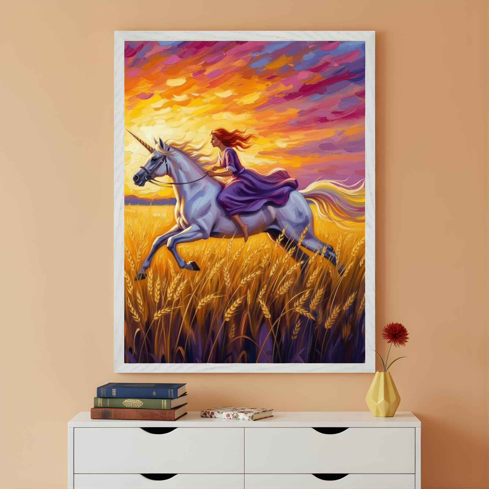 Framed fantasy painting shows a girl in a flowing purple dress riding a white unicorn through a golden wheat field at sunset, with vivid orange, yellow, and magenta clouds filling the sky. The artwork hangs above a white dresser with stacked books and a single red flower in a geometric yellow vase, creating a bold and whimsical focal point in the room.