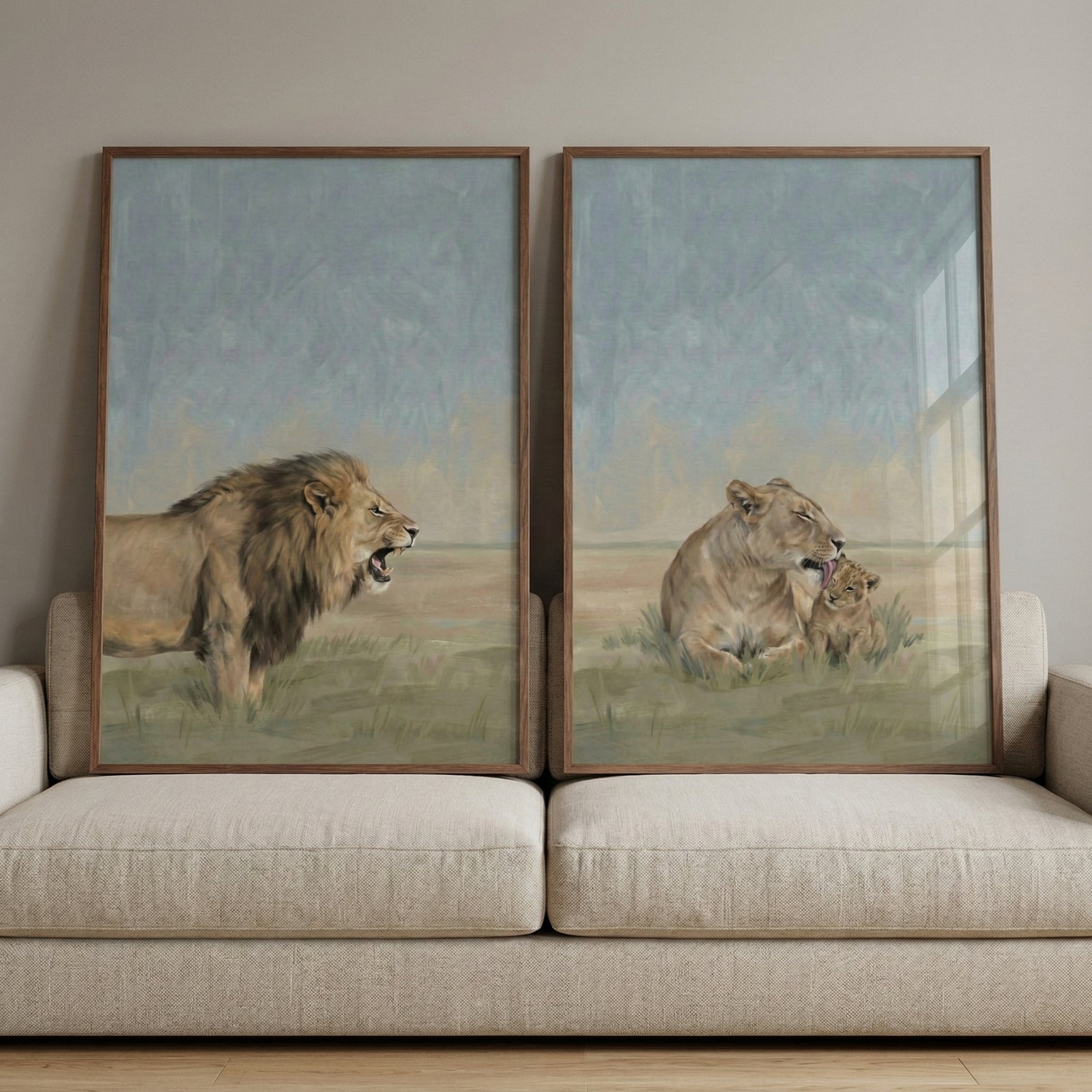 Two framed wildlife paintings lean against a beige sofa. One artwork shows a male lion standing in grassy savanna with its mouth open in a roar while the other shows a lioness gently grooming her small cub in tall grass. The soft neutral tones create a calm nature inspired scene.