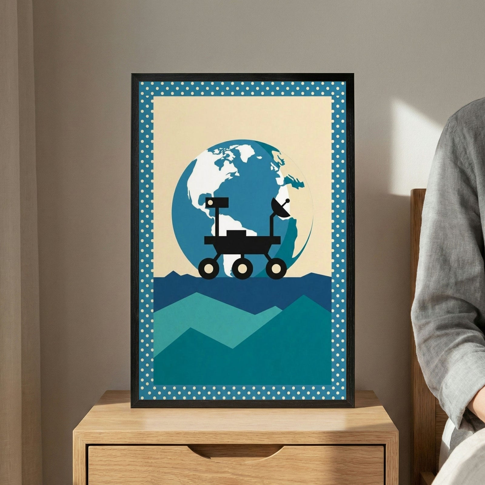 A minimalist graphic illustration in a black frame sits on a wooden nightstand. The artwork depicts a black silhouette of a space rover on a jagged landscape of teal and blue geometric mountains. A large, stylized blue and white Earth dominates the background against a pale, off-white sky. A portion of a person wearing a grey garment is visible on the right side.