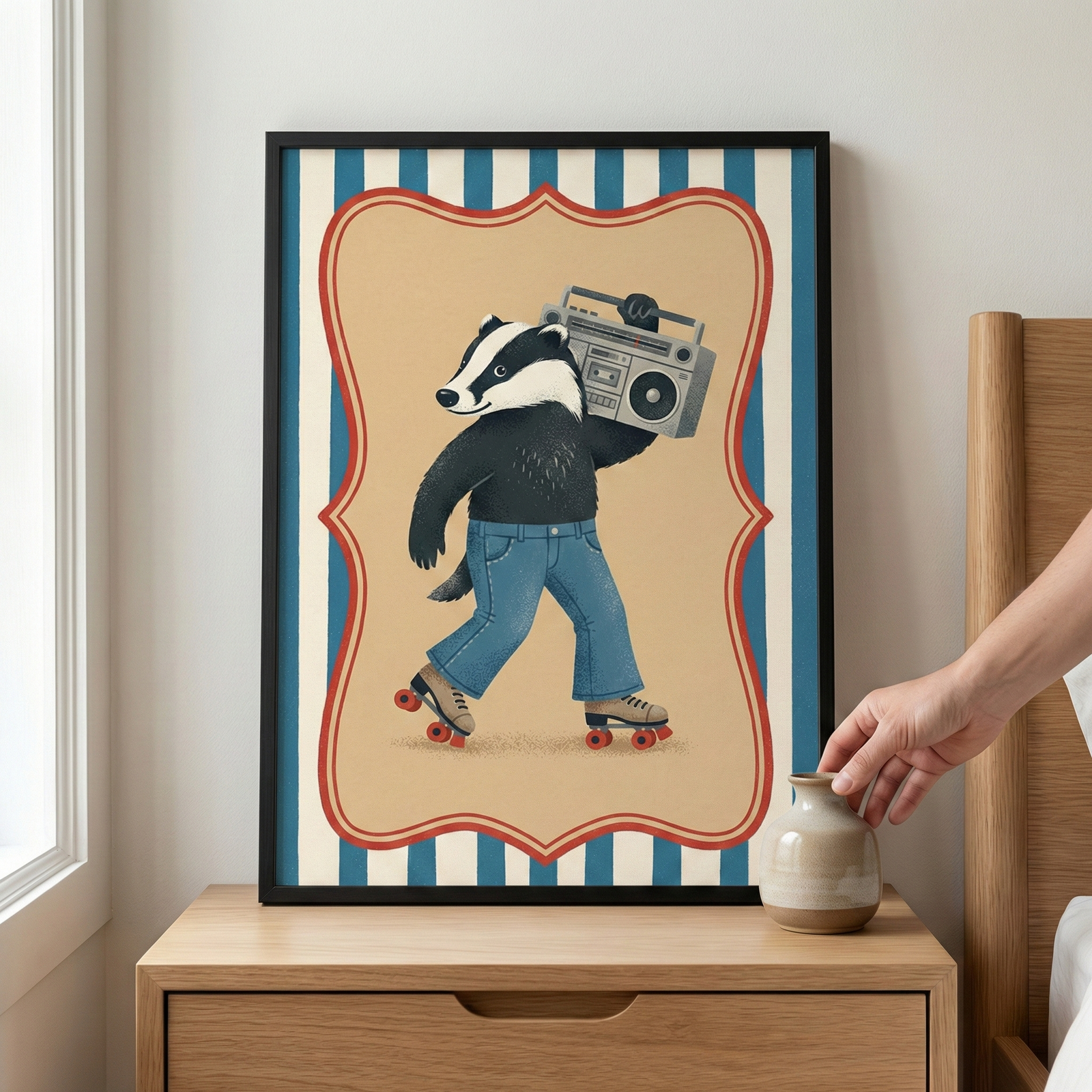 A framed illustration of an anthropomorphic badger sits on a light-colored wooden nightstand. The badger is shown wearing blue denim jeans and tan roller skates with red wheels while carrying a large silver boombox on its shoulder. The character is set against a tan background within a red scalloped border, which is layered over a blue and white vertical striped pattern. A hand is reaching toward a small, neutral-toned ceramic vase placed on the table next to the frame.