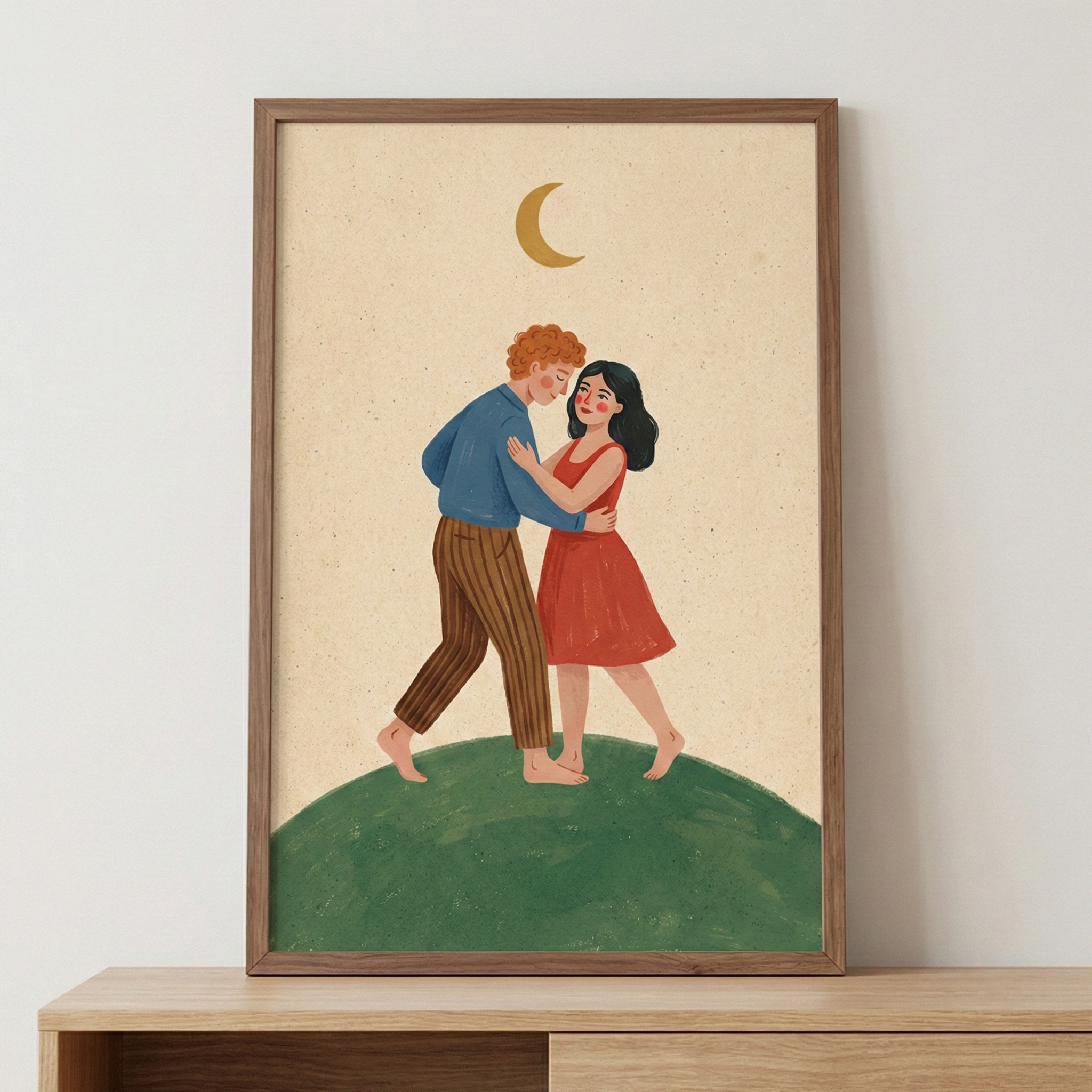 A framed vertical piece of wall art sits on a light wooden surface against a plain white wall. The illustration depicts a man and a woman in a gentle, romantic pose. The man, with curly red hair, wears a blue long-sleeved top and brown vertical-striped trousers. The woman, with black hair, is dressed in a sleeveless red dress. Both are barefoot, standing atop a curved green hill. Above them, a simple golden crescent moon is set against a textured, light beige background. 