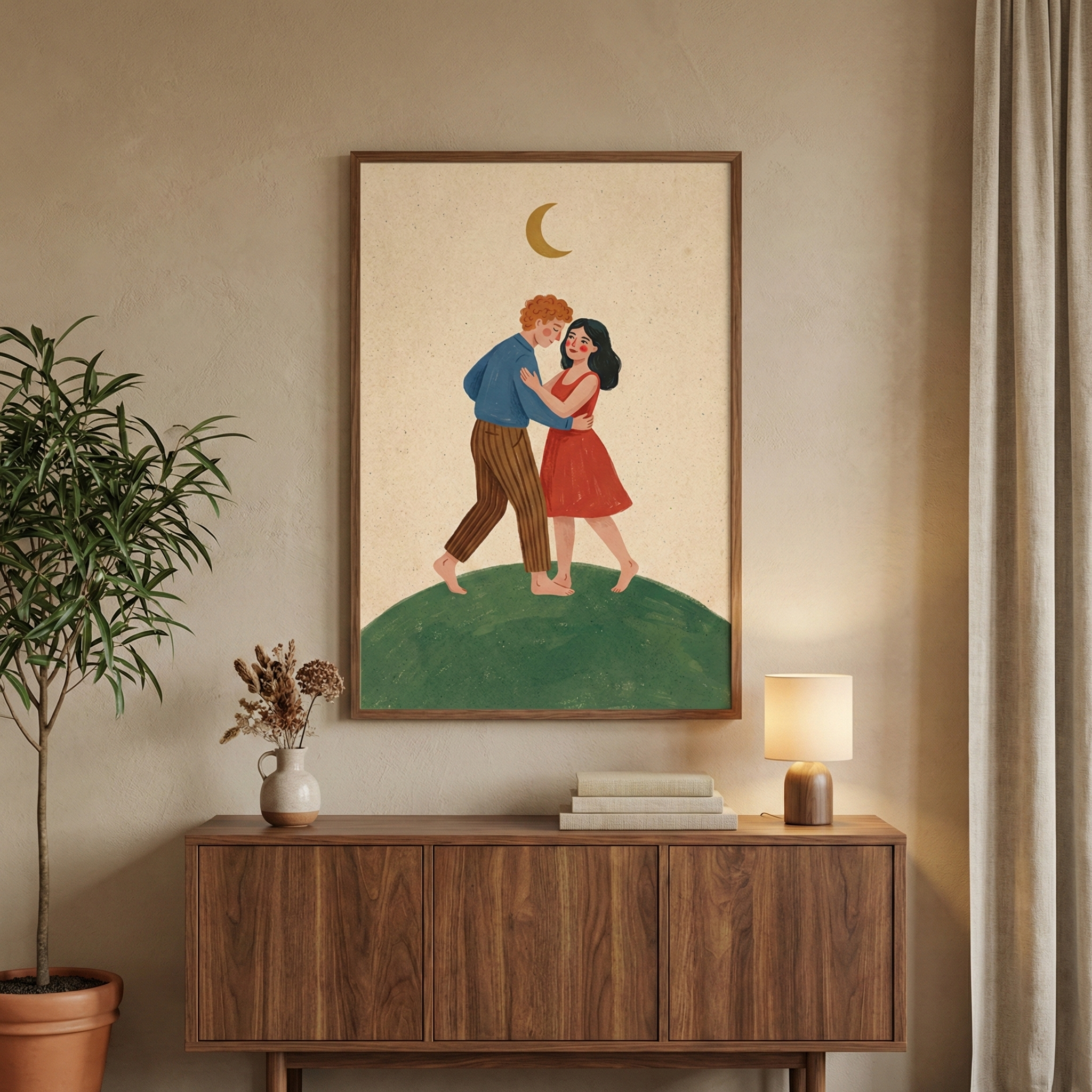 An illustration in a wooden frame hangs on a neutral beige wall. The artwork depicts a couple dancing barefoot atop a rounded green hill under a golden crescent moon. The man has ginger hair and is wearing a blue shirt and brown striped trousers, while the woman has dark hair and wears a red dress. Below the art is a dark wood sideboard. On its surface are a small white vase with dried flowers, a stack of books, and a lamp with a wooden base. To the left is a tall green plant in a pot.