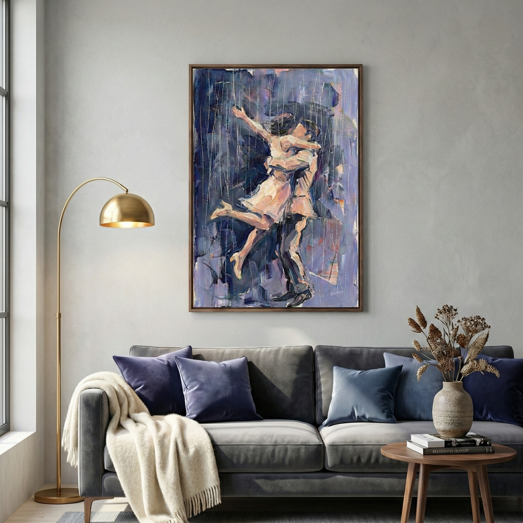 A contemporary living room centers on a vertical framed artwork depicting a couple in an intimate embrace. The art is created in a textured, expressive style with a palette of lavender, indigo, and cream. Below, a dark grey sofa is decorated with velvet pillows in plum and slate blue tones. A chunky cream-colored knit throw is draped over one arm. To the left, a slender gold floor lamp with a dome shade curves gracefully. On the right, a small wooden table holds books and a vase of dried botanicals.