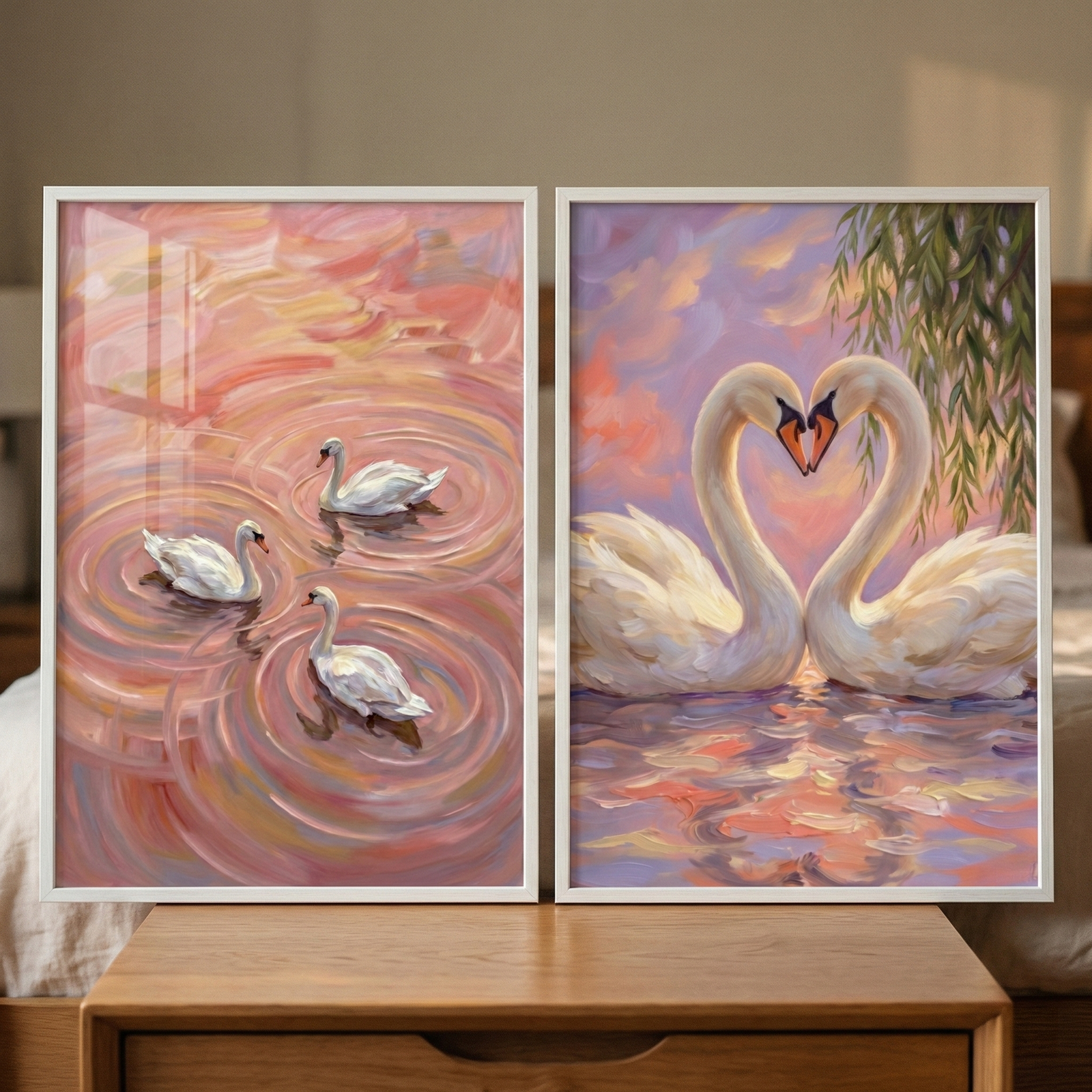 Two framed artworks are displayed side-by-side on a light-colored wooden nightstand. The frame on the left shows three white swans swimming in water rendered in soft shades of rose, peach, and light brown. Concentric circular ripples emanate from each swan, creating a rhythmic pattern across the surface. The frame on the right depicts a pair of white swans floating on calm water, their long necks curved toward each other to form a heart shape.