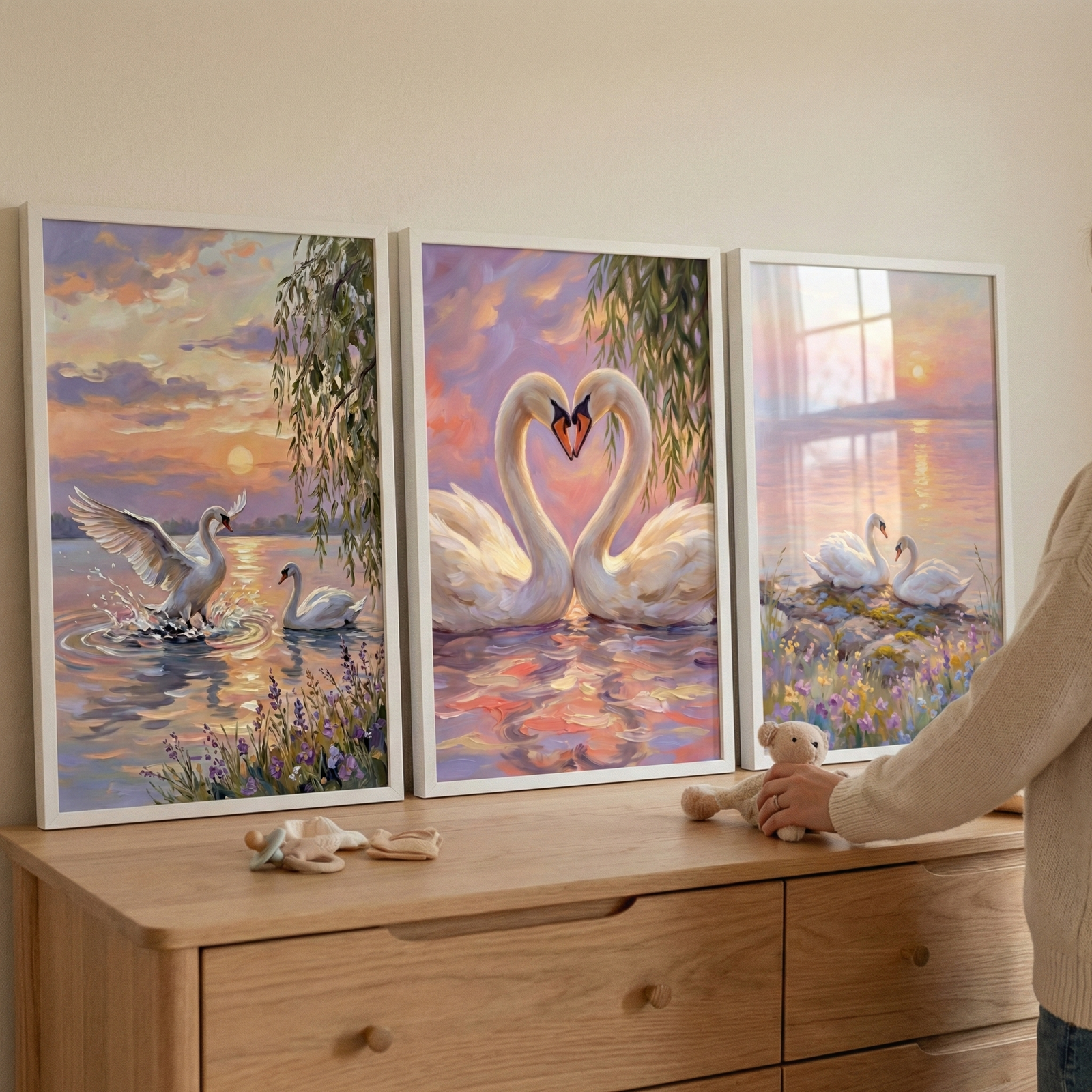 Three framed pieces of swan-themed artwork are displayed side-by-side on a light-toned wooden dresser against a plain wall. The left image depicts a swan with its wings spread, landing in water with a splash during a sunset, while another swan swims in the background. The center image features two swans with their necks curved toward each other to create a heart shape, framed by drooping willow branches against a pastel sky. 