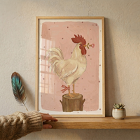 A wooden-framed art print leans against a beige wall on a wooden shelf. The illustration depicts a white rooster with a red comb and wattle standing on a tree stump. It holds a small pink flower in its beak, and a red-and-white checkered ribbon is tied around its leg. The background is soft pink with tiny star shapes. To the left, a large iridescent feather leans against the wall, while a small succulent in a white pot sits on the right. A hand in a knit sweater touches the shelf edge.