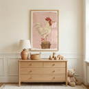 A cozy, neutral-toned child's room features a light-wood framed illustration hanging on a beige wall with decorative wainscoting. The artwork depicts a white rooster standing on a brown wooden stump against a dusty pink background. The rooster has a red comb, a small pink flower in its beak, and a thin red ribbon tied around one leg. Below the artwork is a light-colored wooden dresser with four drawers.