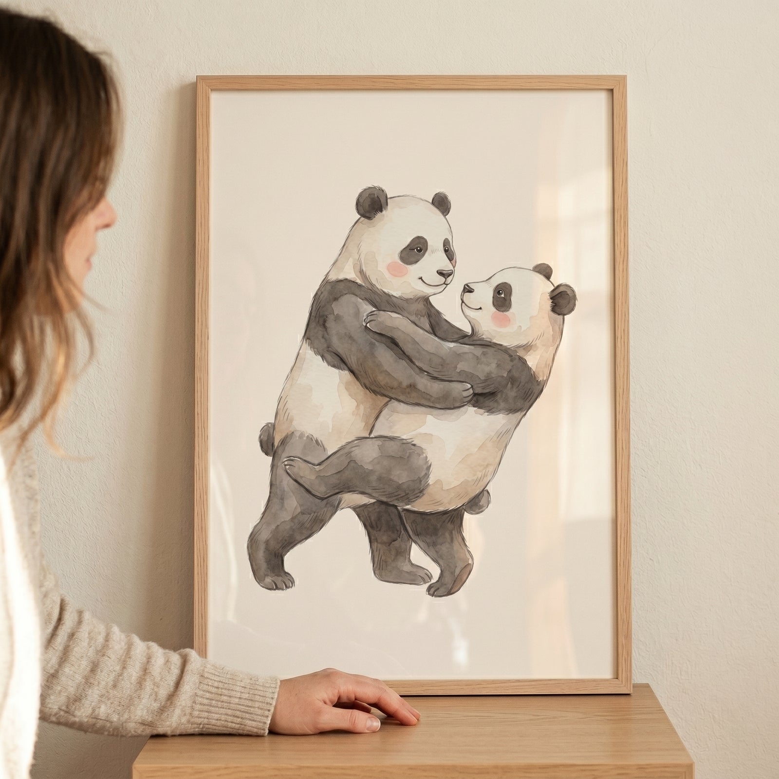 Two pandas embrace in a framed rosy cheek dancing panda artwork on a wooden table beside a woman.