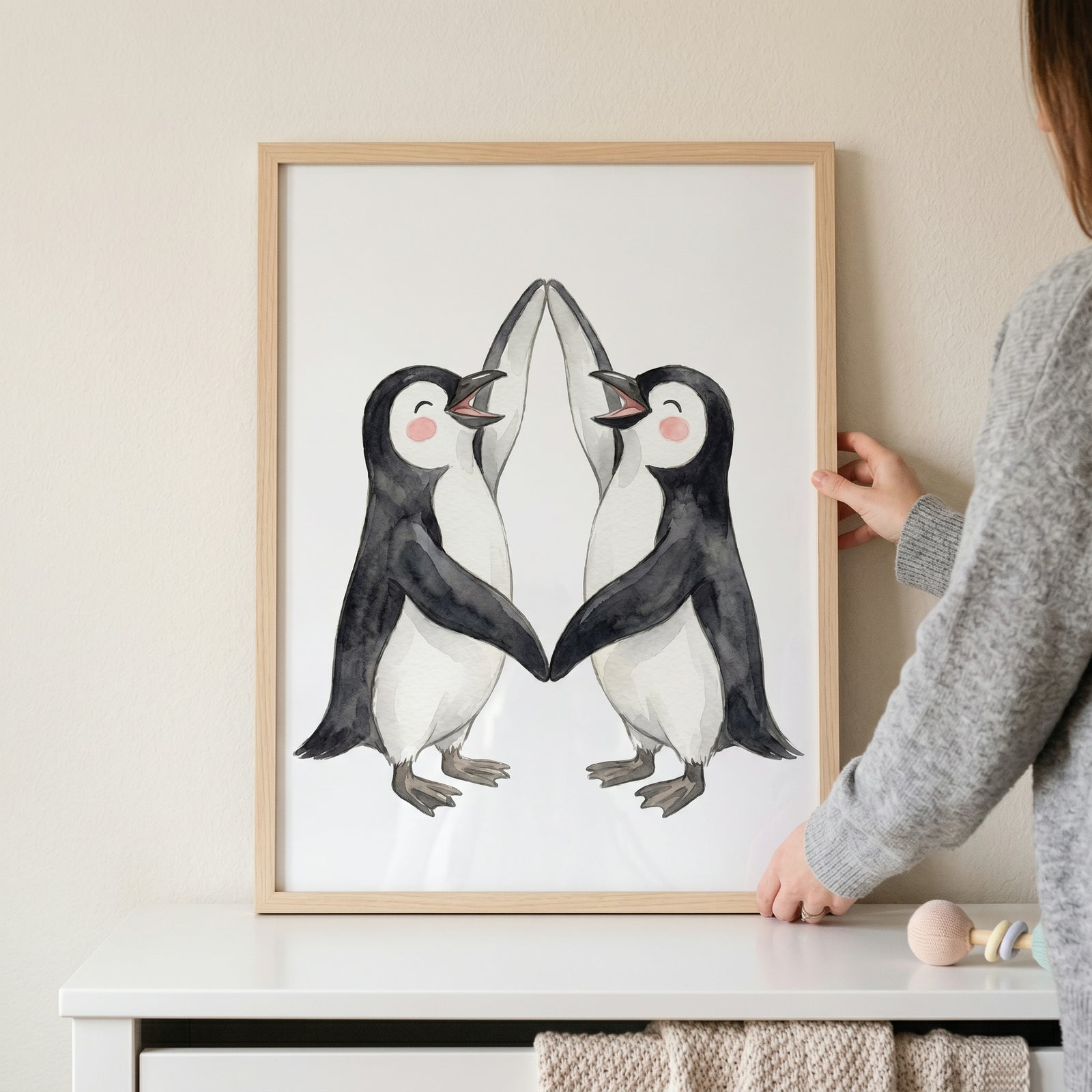 Two watercolor penguins touch flippers in this rosy cheek dancing penguin wall art held above a white dresser.