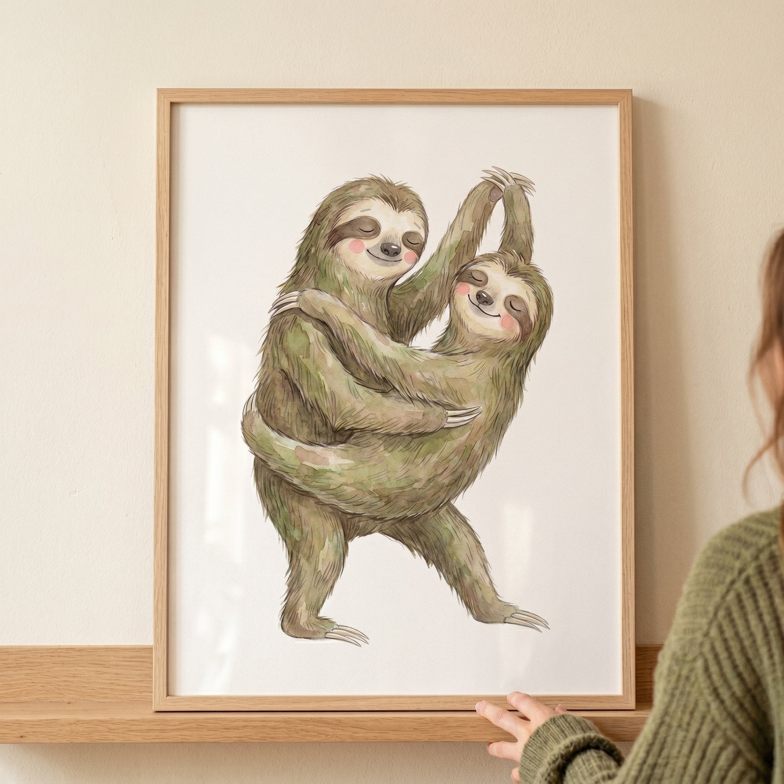 Two sloths embrace in this rosy cheek dancing sloth print, displayed in a light wood frame resting on a shelf.