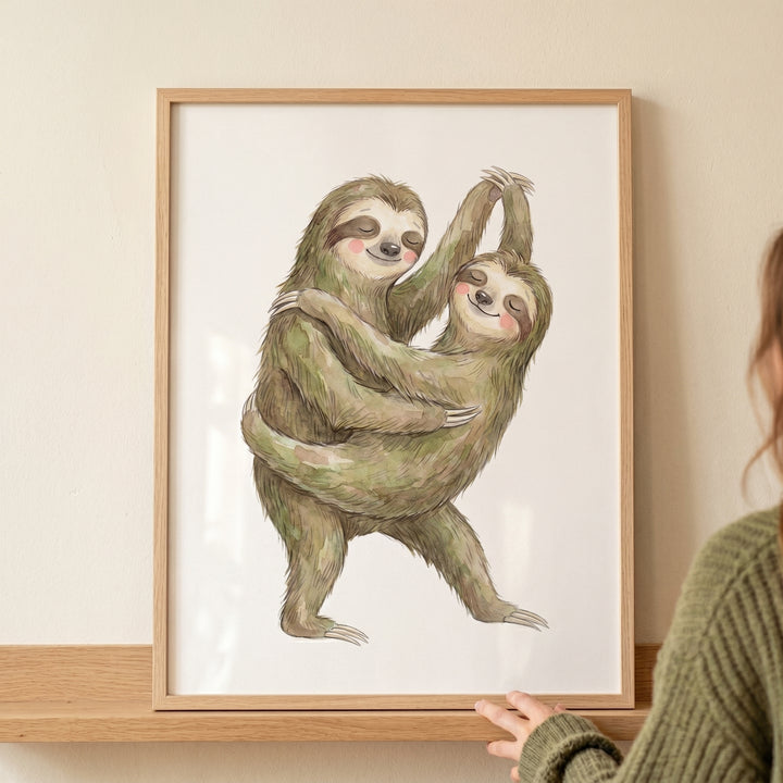Two sloths embrace in this rosy cheek dancing sloth print, displayed in a light wood frame resting on a shelf.