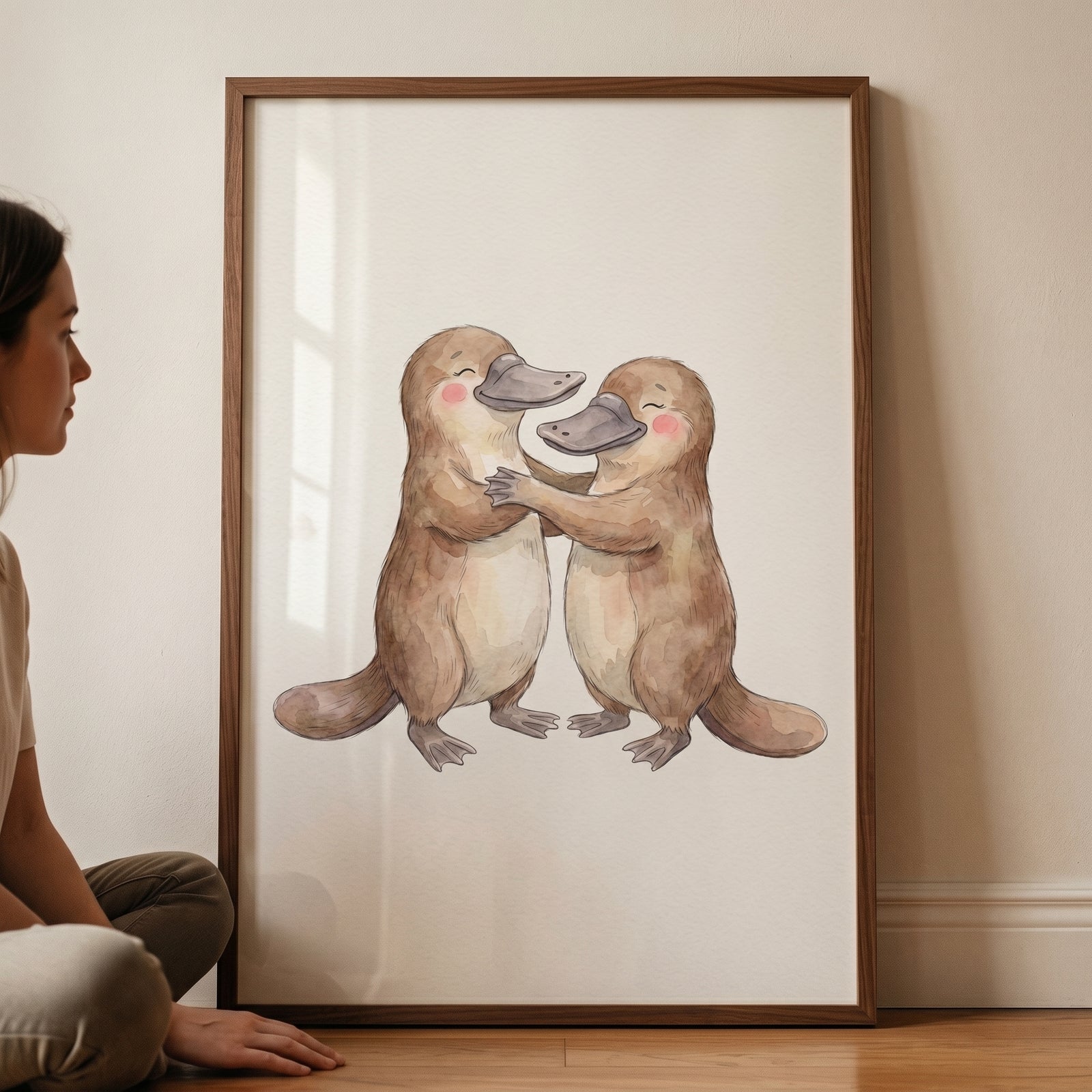 Two cute watercolor platypuses embrace in this framed rosy cheek hugging platypus wall art leaning against a wall.