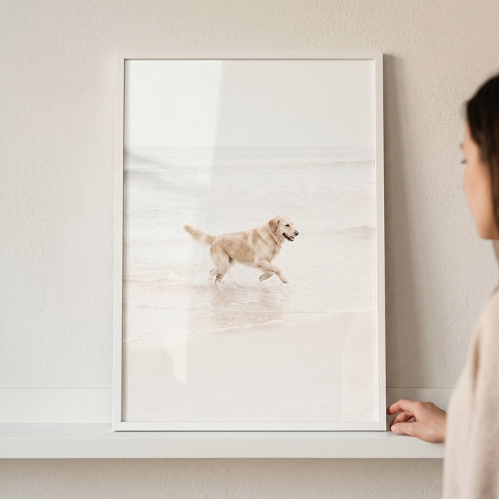 A woman looks at a framed running beach golden retriever wall art piece of a dog splashing in ocean waves.
