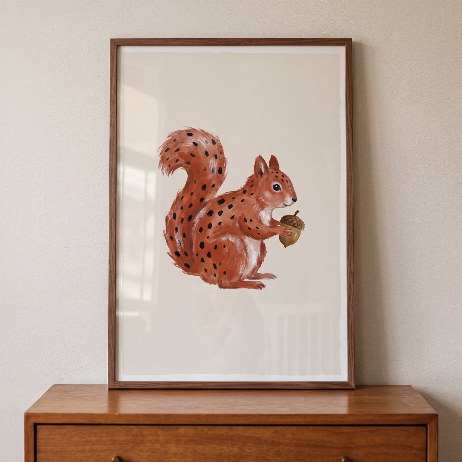 Framed rust spotted acorn squirrel wall art sits on a wooden dresser against a neutral wall.