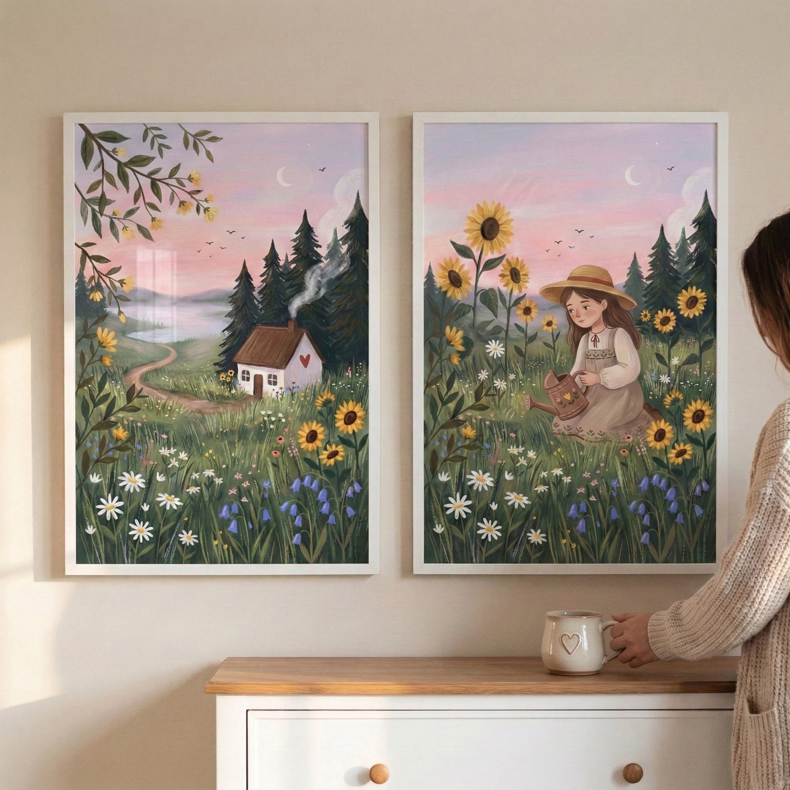 Two framed countryside illustrations hang above a light cabinet while a person holds a mug nearby. One shows a small cottage with smoke rising from the chimney beside a winding path through a wildflower meadow under a pink evening sky with a crescent moon while the other shows a girl in a hat watering flowers among tall sunflowers and daisies in the same soft sunset landscape.