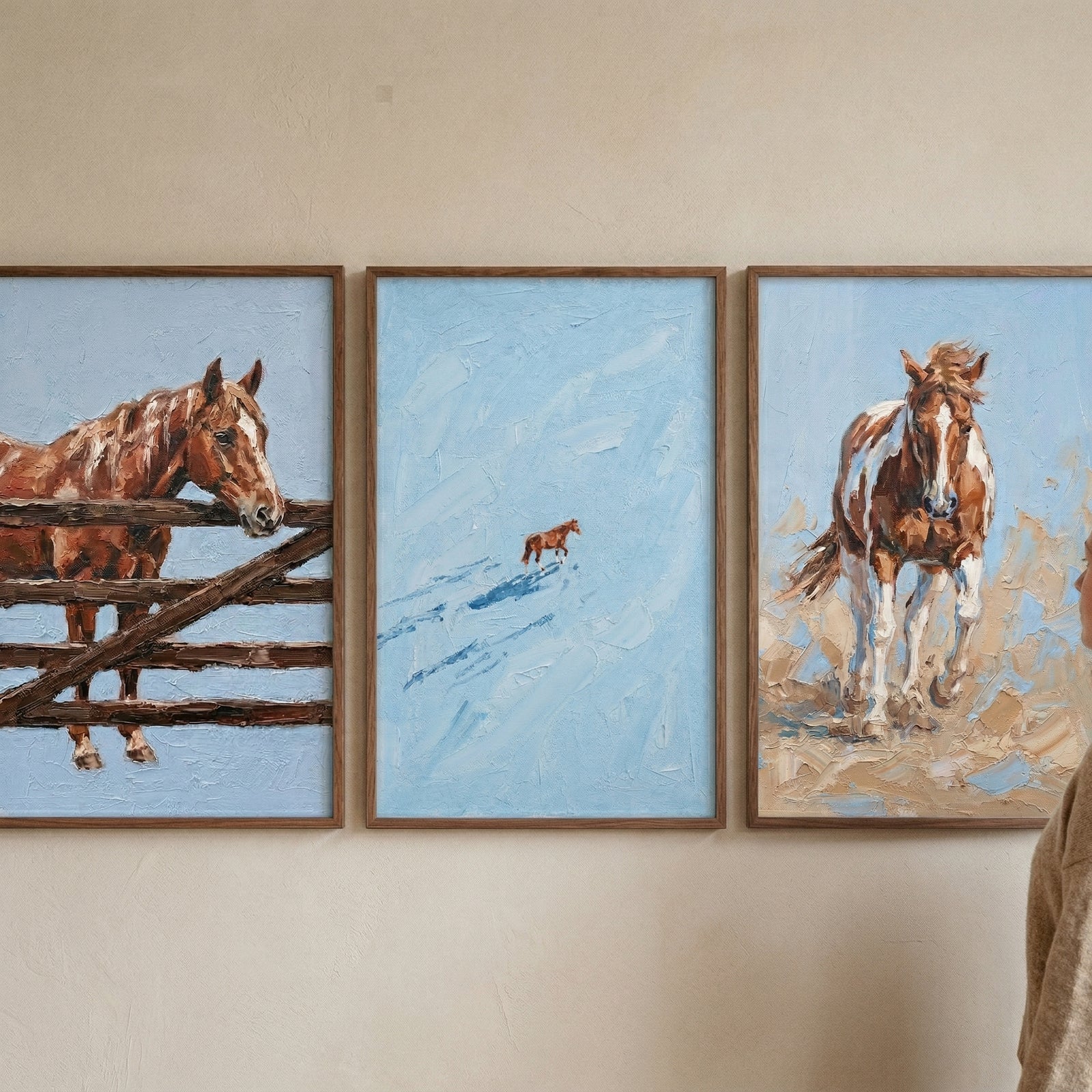 Two framed textured horse paintings displayed on the floor, one showing a close up of a brown horse with a light mane and the other showing a full horse standing on rocky ground against a soft blue background. A person kneels beside the artwork, suggesting placement in a warm rustic interior.