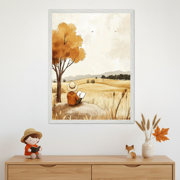 Framed wall art featuring an autumn-toned illustration of a child in a wide-brimmed hat sitting under a tree and sketching in a notebook, overlooking golden fields and distant hills. Below the artwork sits a dresser with a matching plush doll reading a book, a wooden fox figure, and a vase with wheat stalks and an orange maple leaf.