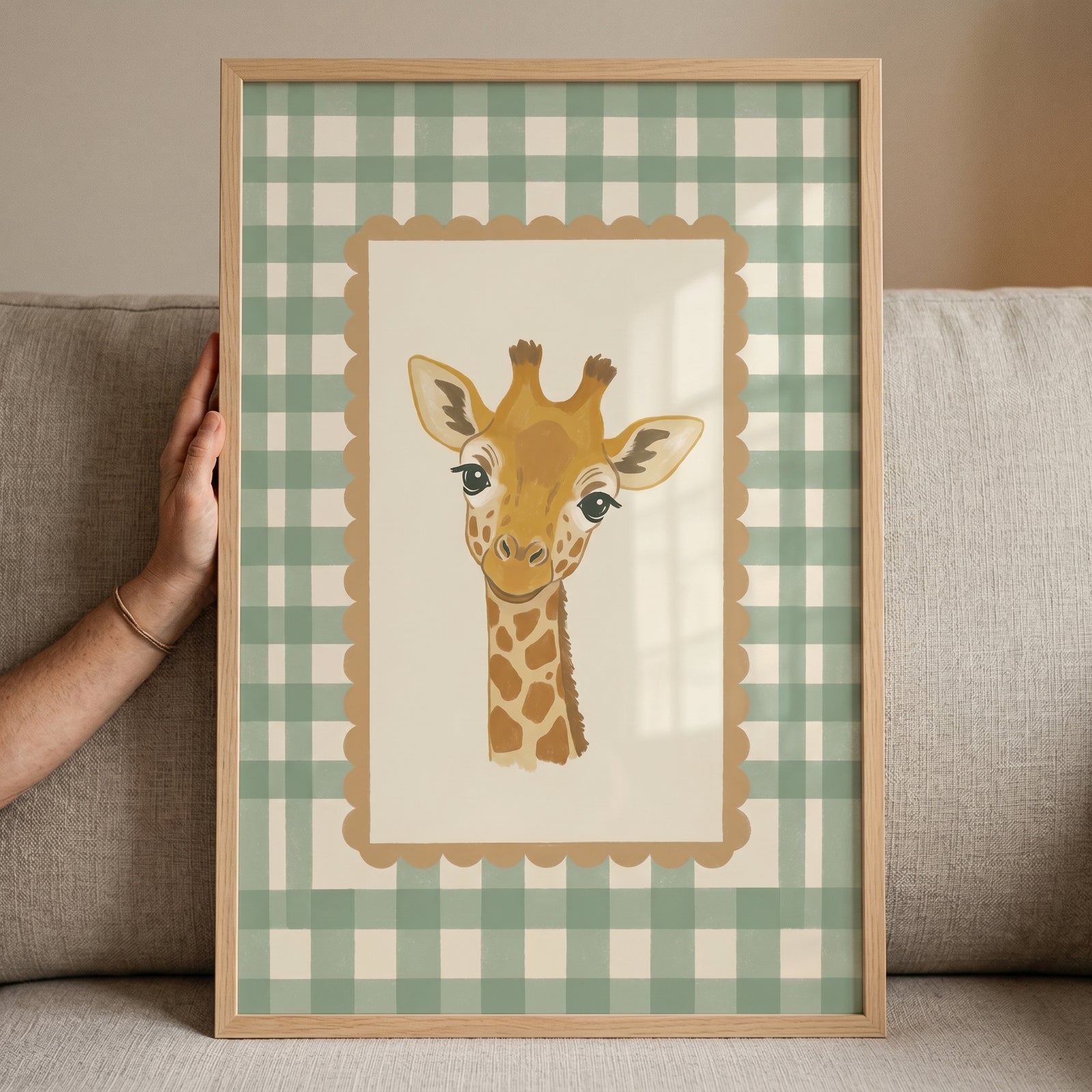 Framed sage green gingham giraffe wall art with a scalloped border held by a hand against a gray couch.