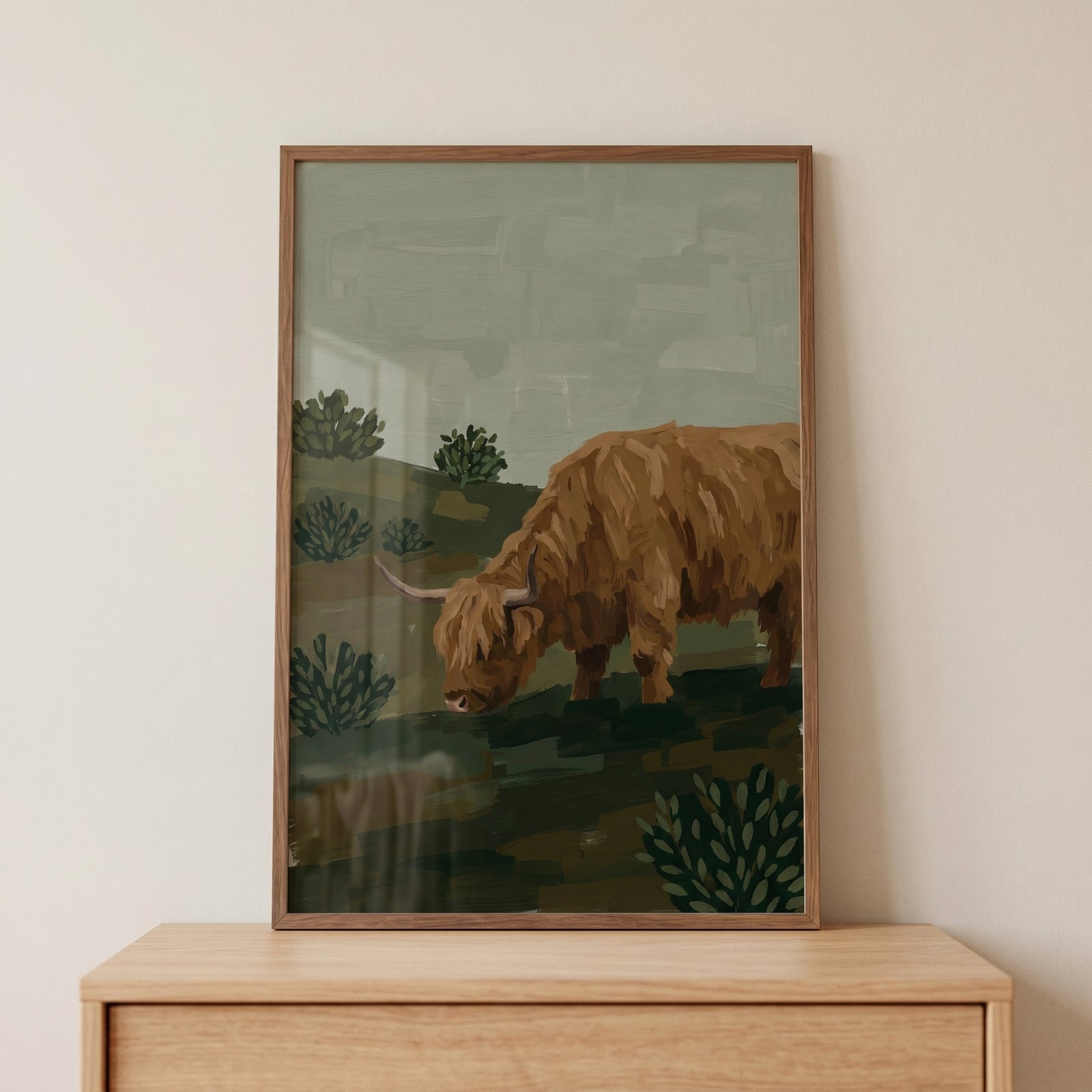 Framed sage green highland cow wall art sits on a wood dresser, featuring a shaggy cow in a muted landscape.