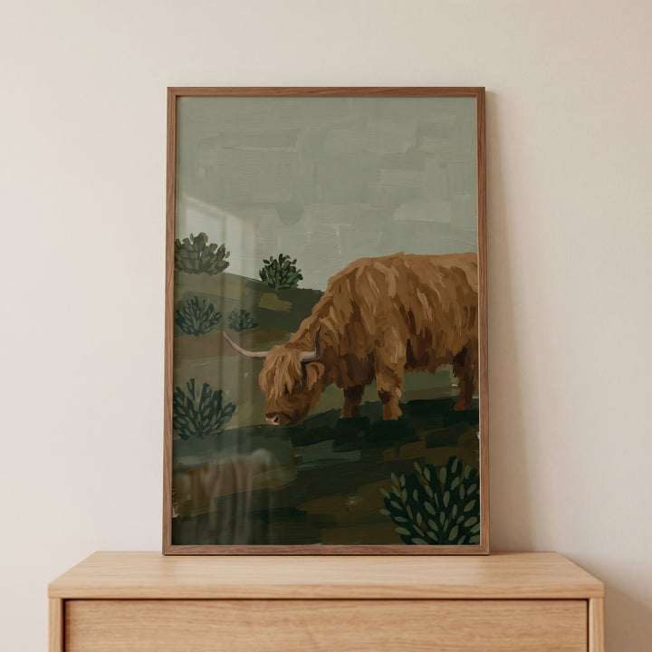 Framed sage green highland cow wall art sits on a wood dresser, featuring a shaggy cow in a muted landscape.