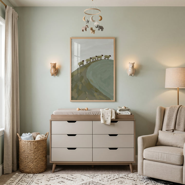 Sage green hill sheep wall art above a beige dresser in a nursery with a mobile, woven basket, and rocker chair.