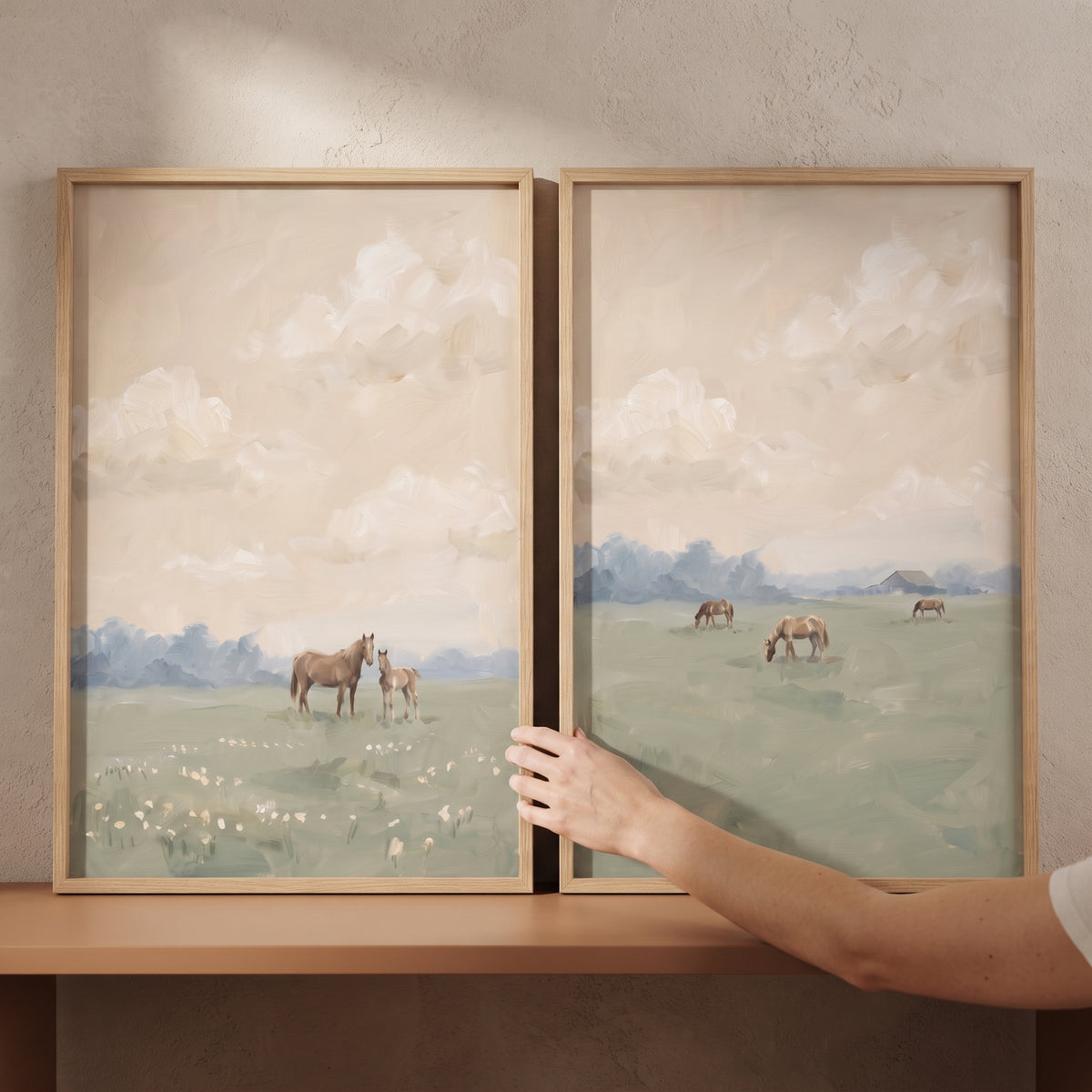 A hand touches a sage green meadow horse wall art set of 2 depicting horses in a hazy field under a soft beige sky.