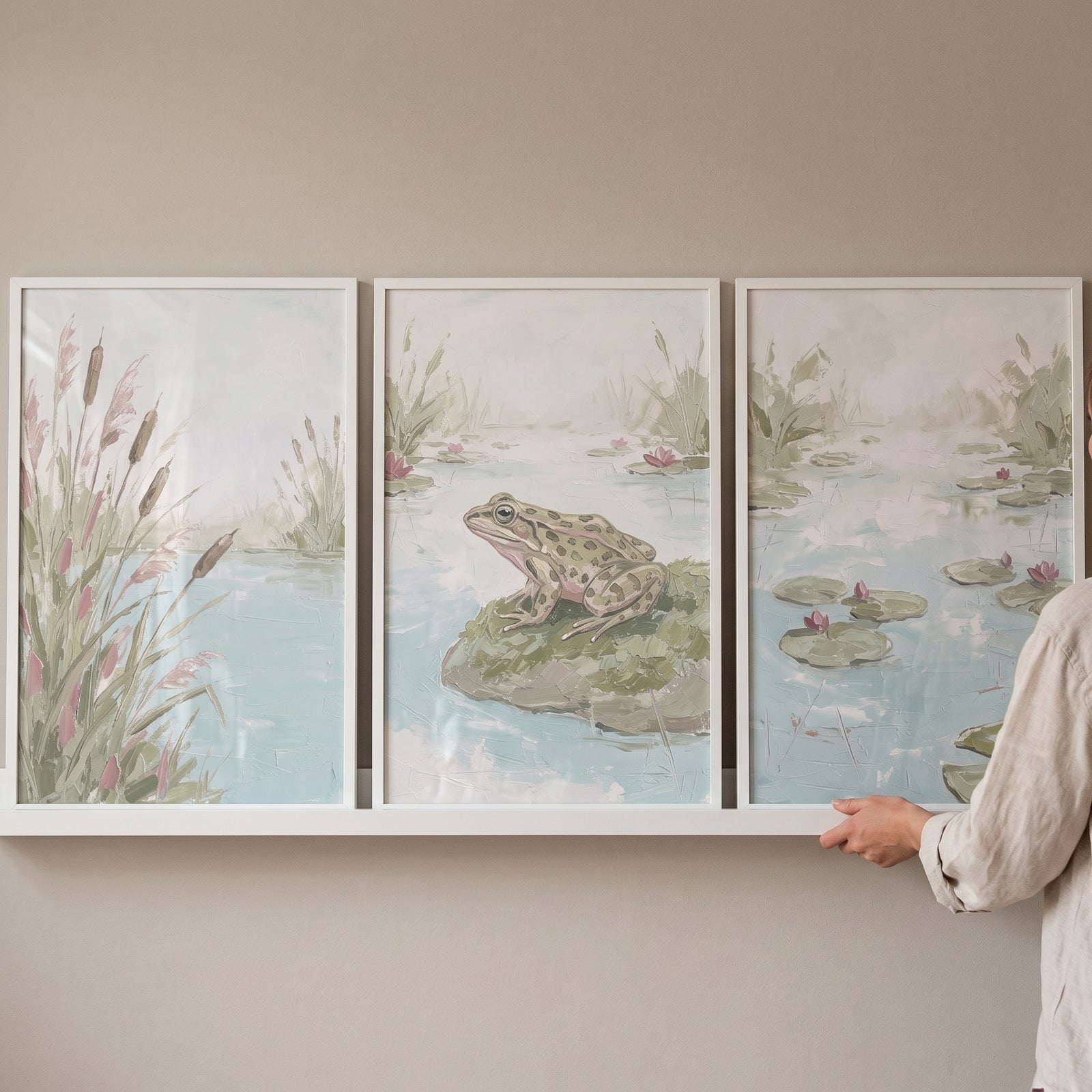 Sage green nature pond wall art set of 3 featuring a frog, reeds, and water lilies in white frames on a ledge.