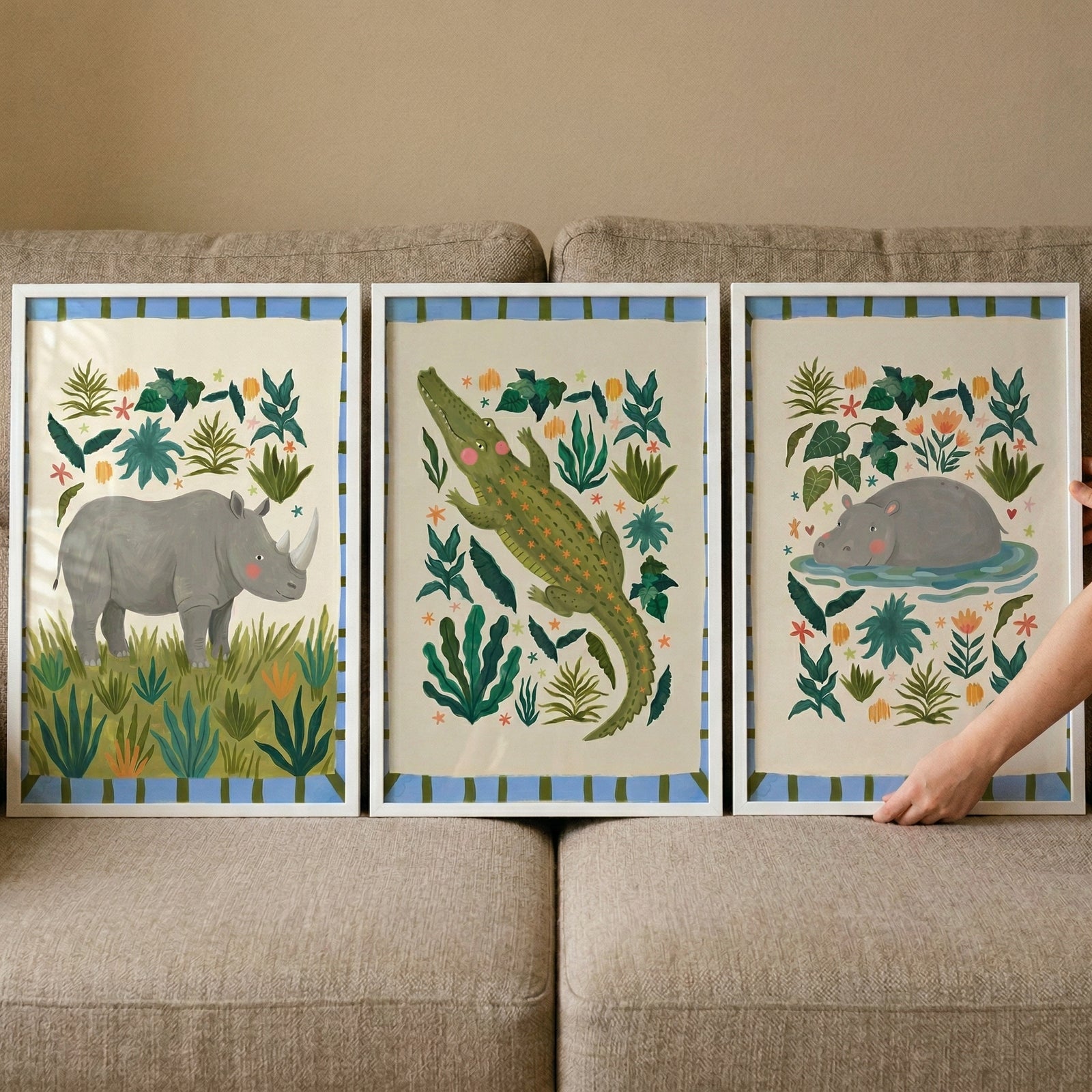 Sage green safari animal wall art set of 3 features rhino, crocodile, and hippo illustrations in white frames on a sofa.