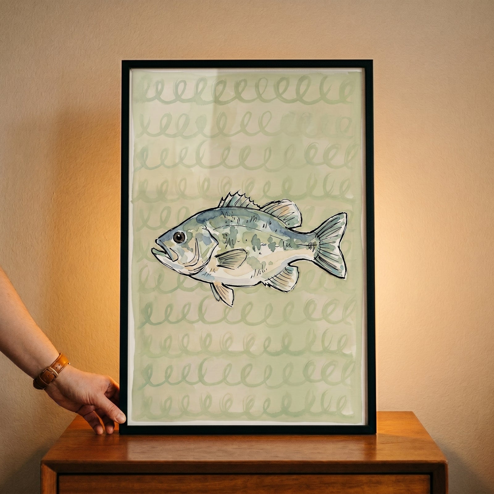 Sage green spiral bass fish wall art in a black frame sits on a wooden table with a hand touching the corner.