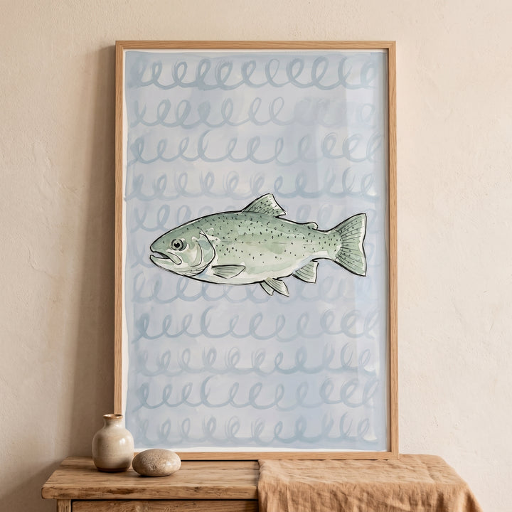 Sage green spotted trout fish wall art in a wooden frame displayed on a wooden table with small ceramic objects.