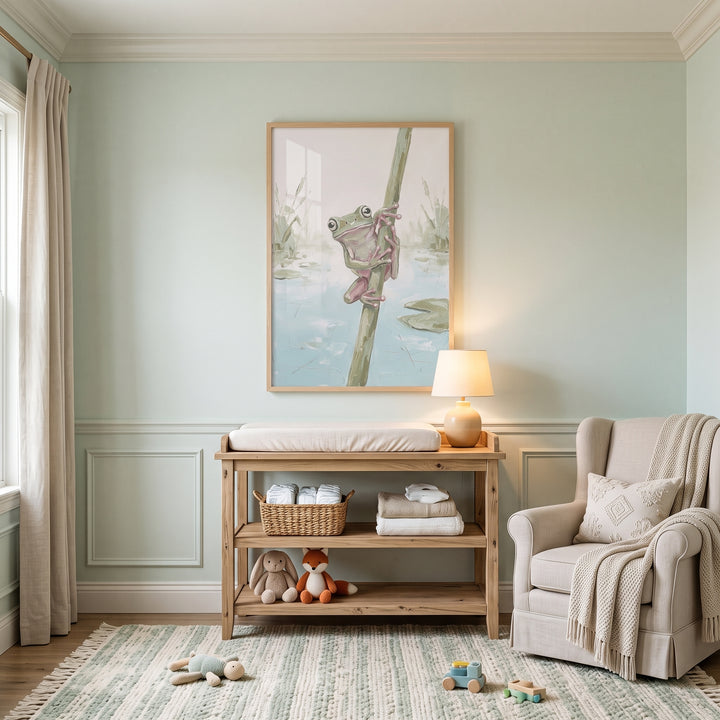 Sage green tree frog wall art hangs above a wooden changing table in a nursery with an armchair and a striped rug.