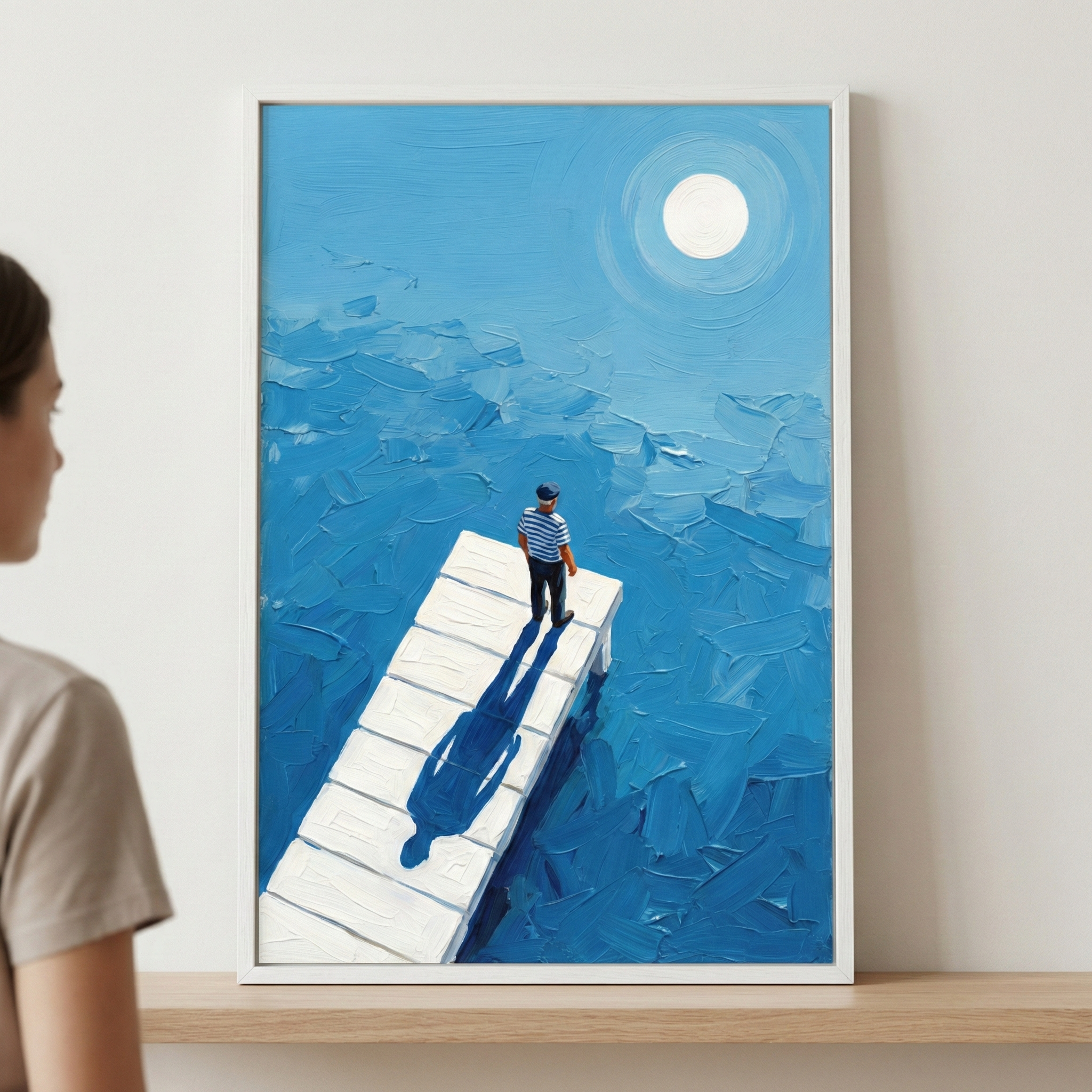 A vertical framed artwork depicts a person in a blue and white striped shirt standing on a white wooden dock that extends into a vast sea of blue. The water and sky are rendered with thick, textured strokes in varying shades of blue. A large white circle glows in the upper portion, casting a long, dark blue shadow of the person onto the dock. The piece is held in a simple white frame and rests on a light wooden shelf. A person is partially visible on the left side of the frame, looking at the display.