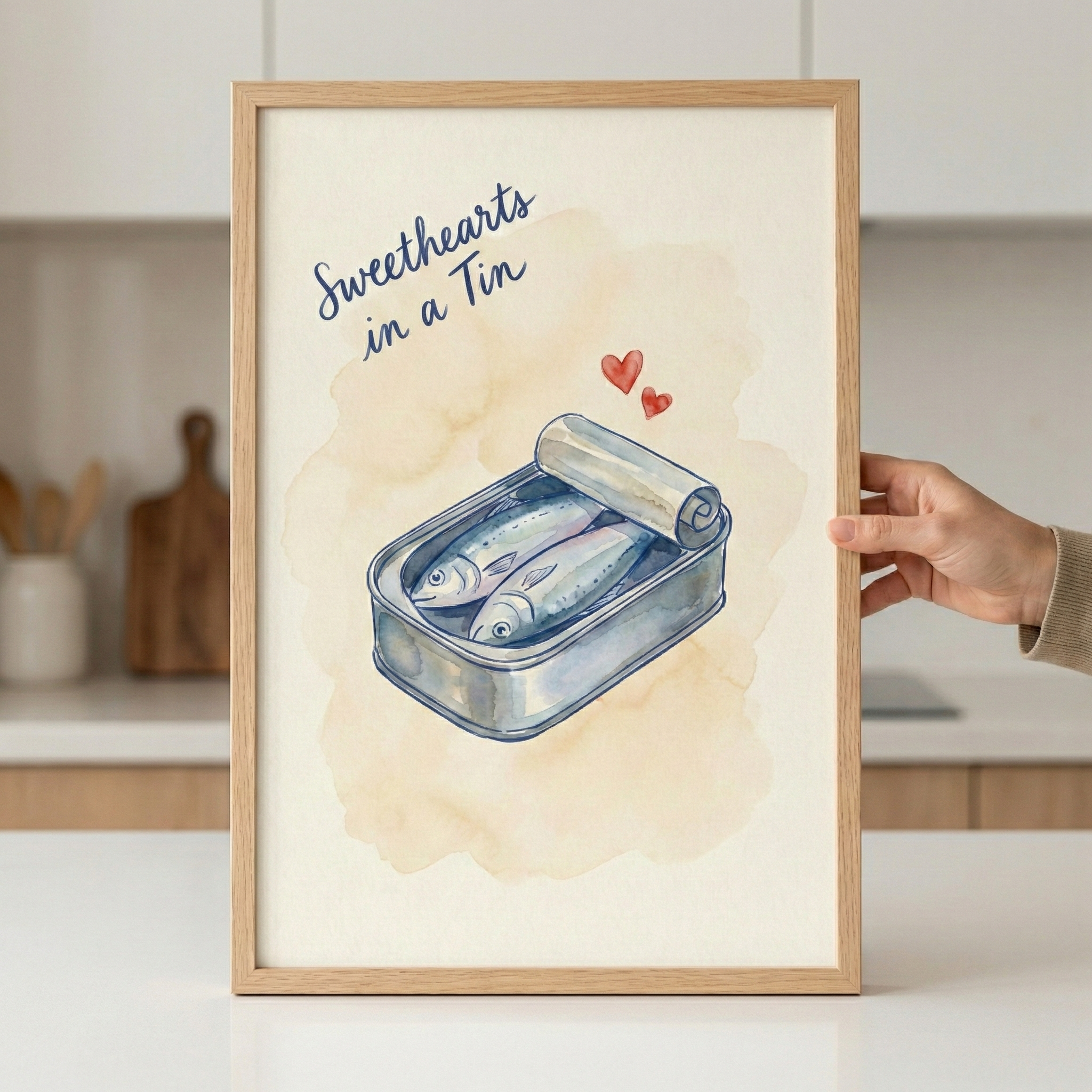A light wood-framed piece of art held by a hand, showing a watercolor illustration of two small fish nestled together in an open metal tin. Above the fish are two small red hearts. The background of the illustration has a soft peach wash, and the words "Sweethearts in a Tin" are written in a dark blue script in the upper left corner. The framed artwork is being held in a bright, modern kitchen.