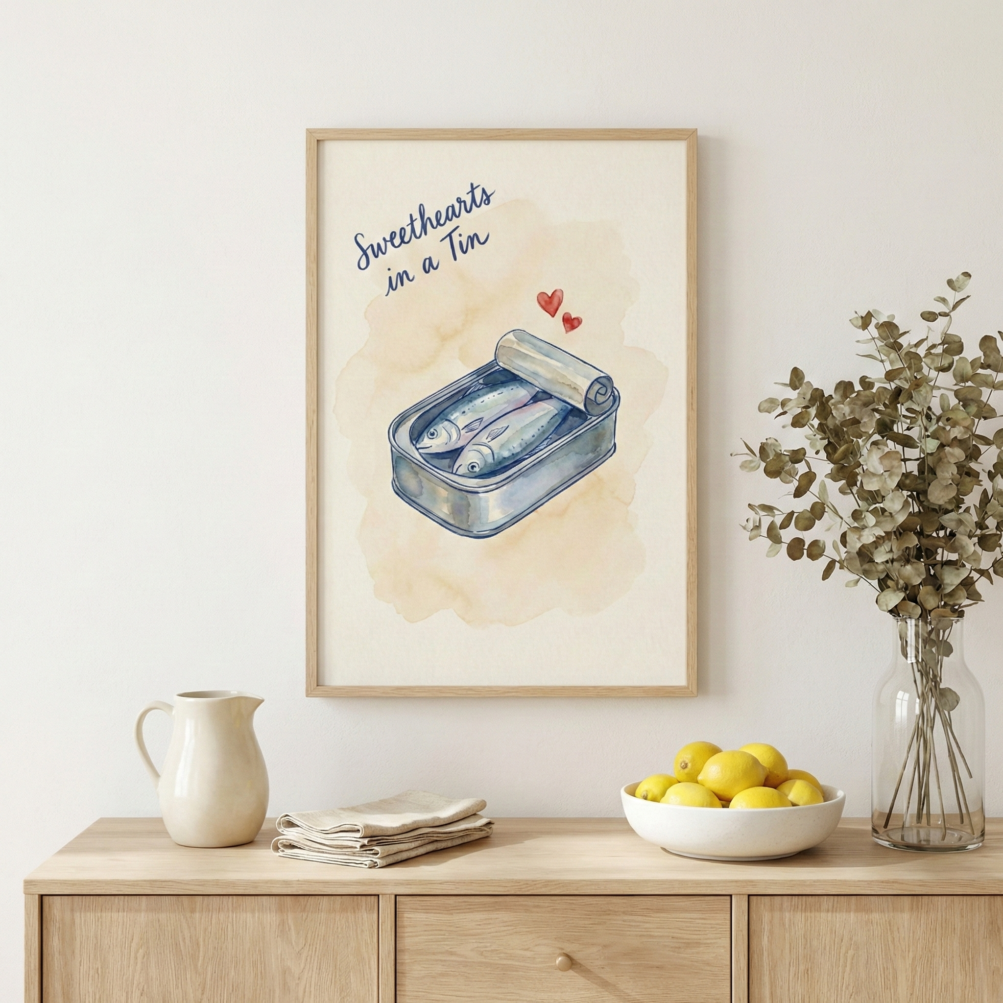 A framed watercolor illustration hangs on a neutral wall above a light wood sideboard. The artwork depicts an open tin of two sardines with two small red hearts floating above them and the cursive text "Sweethearts in a Tin" at the top. On the cabinet below, there's a white ceramic pitcher, a stack of folded napkins, a bowl of bright yellow lemons, and a glass vase with dried eucalyptus sprigs.