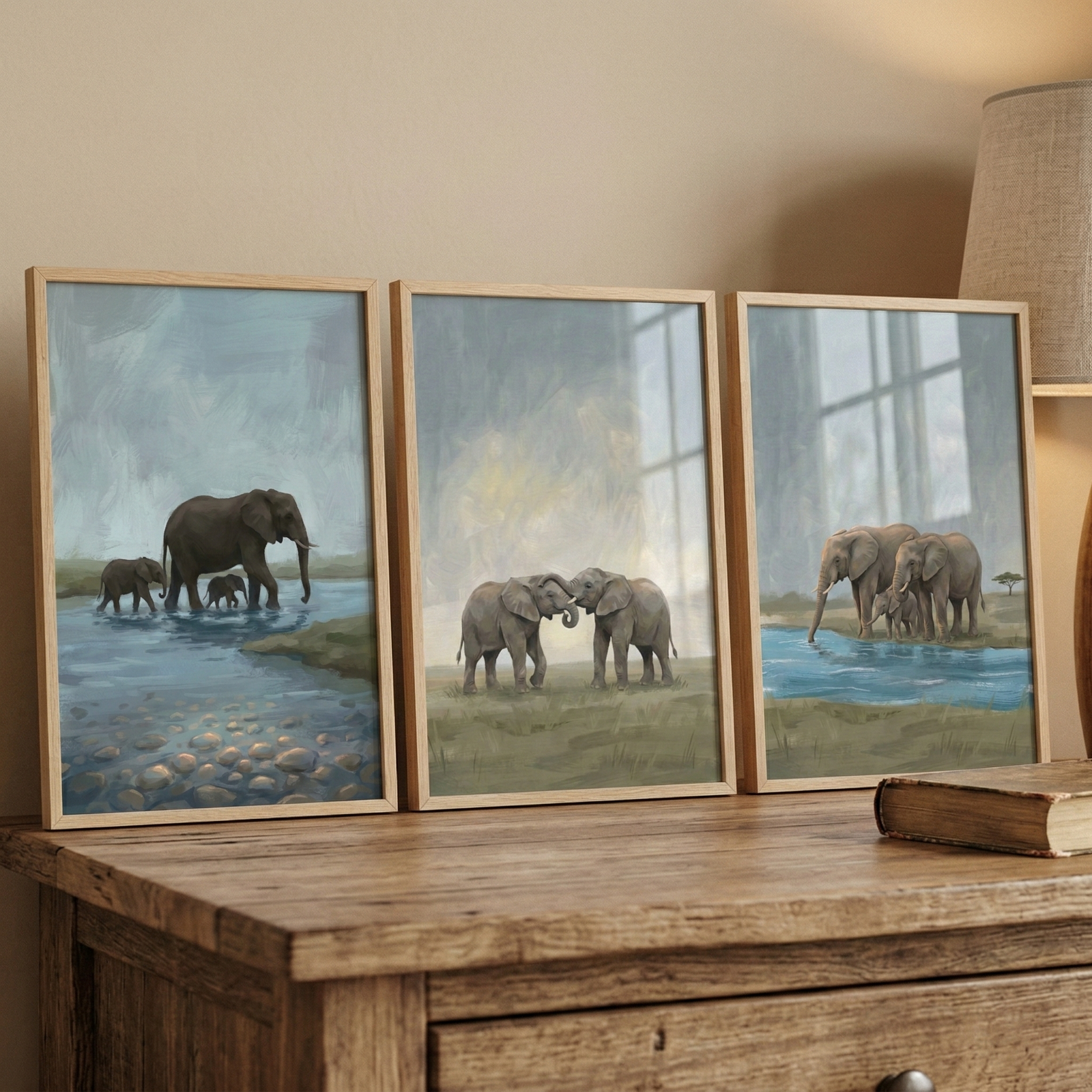 Three framed elephant paintings are displayed on a wooden table beneath a lamp in a warm neutral room. The left painting shows an adult elephant with two calves walking through shallow water, the center painting shows two elephants touching trunks on grassy land, and the right painting shows two adult elephants with a calf beside a blue water edge.