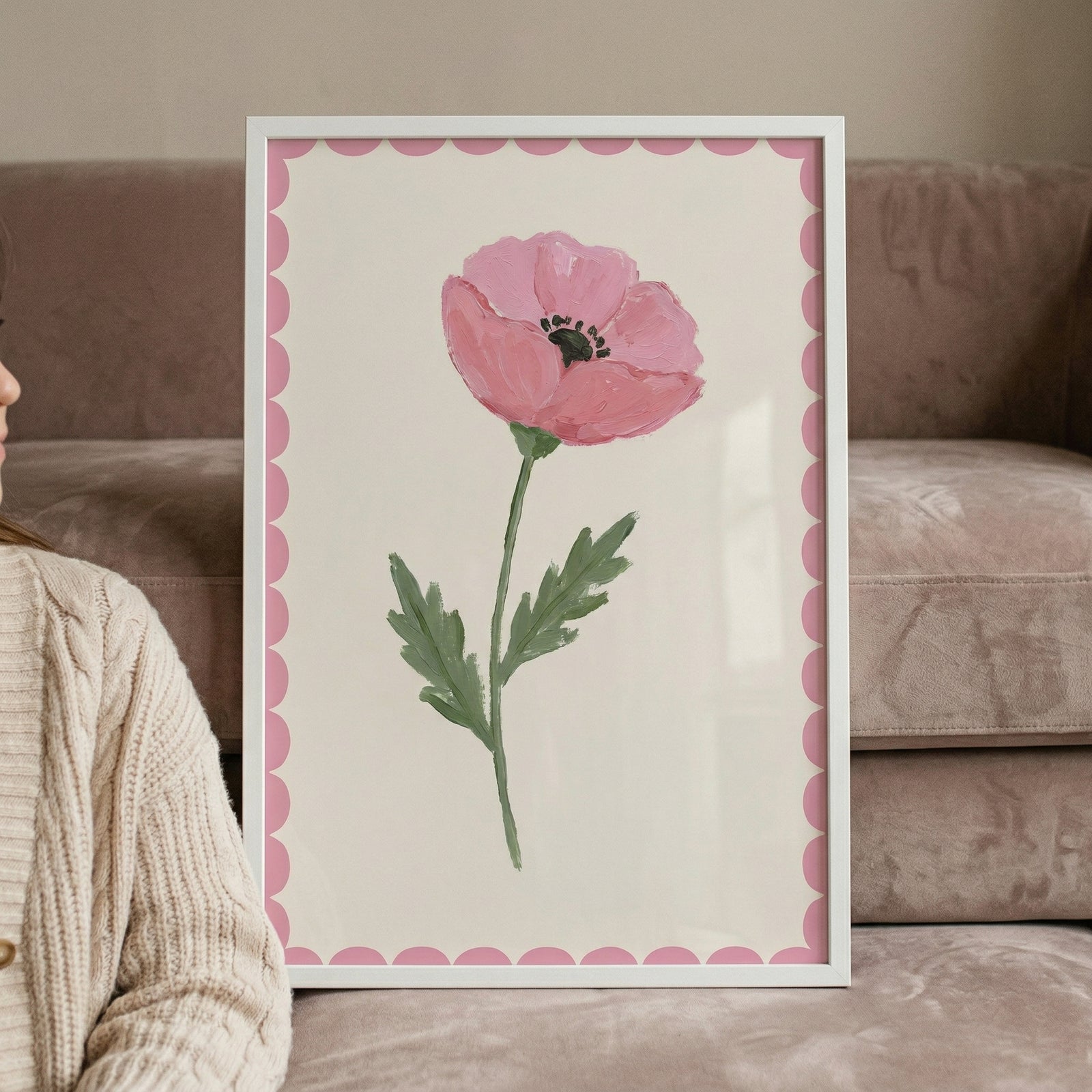 A minimalist framed artwork depicts a single, soft pink flower with a dark center, a green stem, and jagged leaves. The floral illustration is centered on an off-white background, framed by a decorative pink scalloped border and a slim white frame. The framed piece leans against a plush, tan-colored sofa. In the foreground, a person's shoulder and arm are visible, clad in a thick, cream-toned cable-knit sweater, creating a cozy and modern interior scene.