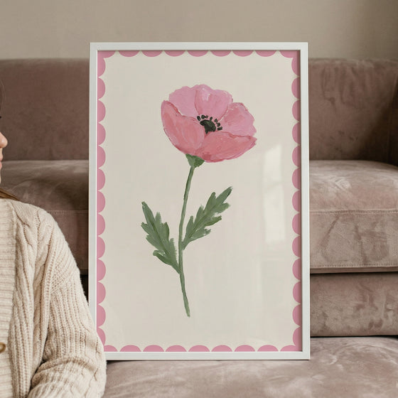 A minimalist framed artwork depicts a single, soft pink flower with a dark center, a green stem, and jagged leaves. The floral illustration is centered on an off-white background, framed by a decorative pink scalloped border and a slim white frame. The framed piece leans against a plush, tan-colored sofa. In the foreground, a person's shoulder and arm are visible, clad in a thick, cream-toned cable-knit sweater, creating a cozy and modern interior scene.