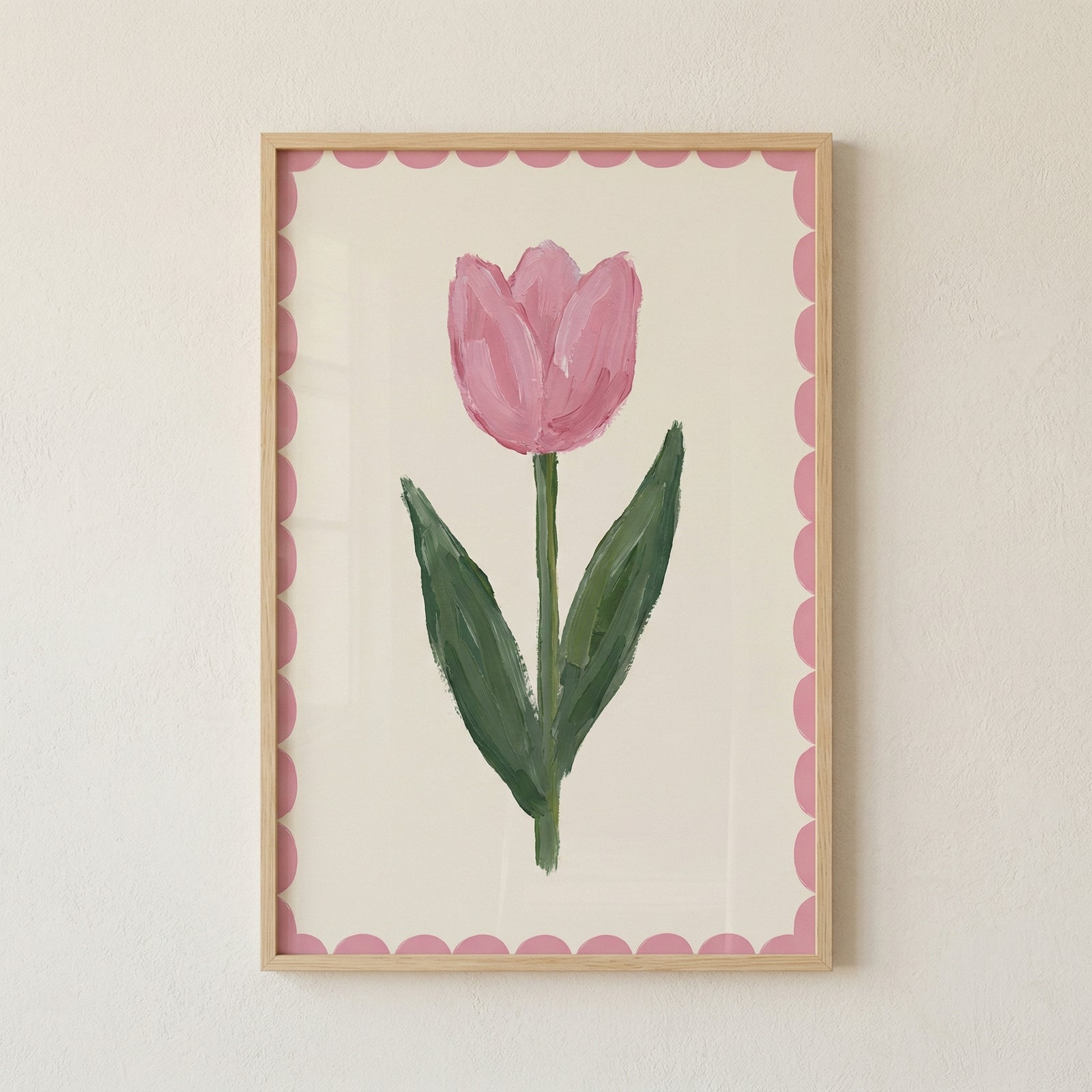 A minimalist floral illustration of a single pink tulip flower. The blossom is rendered with soft pink tones and visible, textured brushstrokes that give it a hand-drawn feel. A slender green stem and two large green leaves extend upwards, also showing expressive strokes and various shades of green. The flower is centered on a light cream background, surrounded by a decorative pink scalloped border. The entire art piece is set within a light-colored wooden frame and hung against a plain, off-white wall.