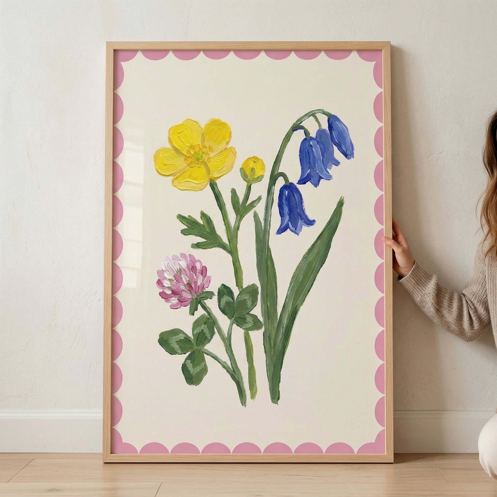 Framed botanical painting of a yellow flower, blue bell shaped flowers, and a pink clover style bloom with green leaves, set against a soft background with a pink scalloped border. A person’s hand gently holds the frame, showing scale and how the colorful floral artwork can be styled in a home.