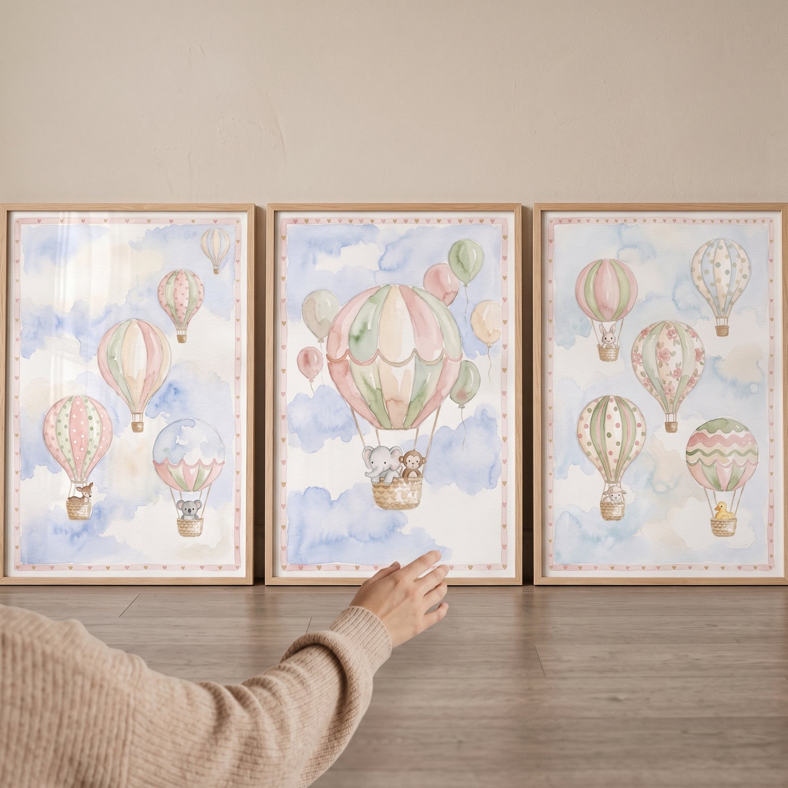 A hand reaches for a scandi watercolor balloon wall art set of 3 with framed prints of animals in hot air balloons.