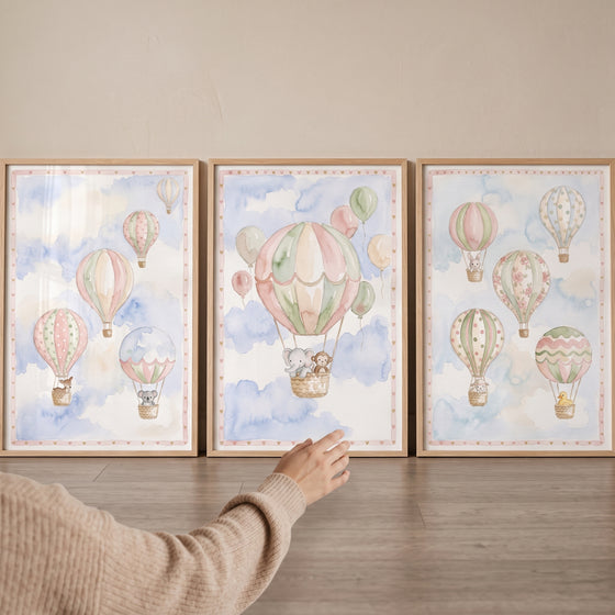 A hand reaches for a scandi watercolor balloon wall art set of 3 with framed prints of animals in hot air balloons.