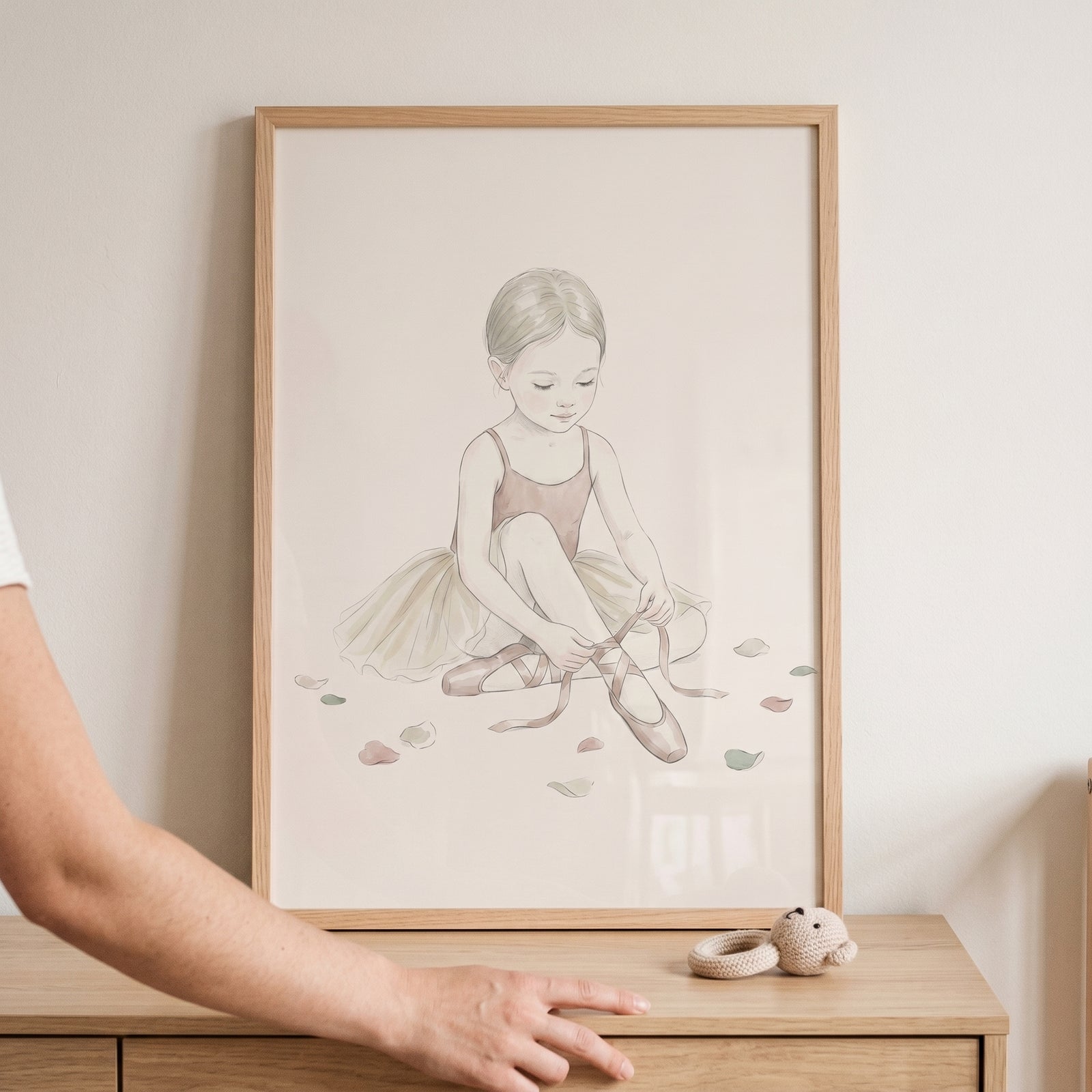 Framed scattered petal seated ballerina wall art on a light wood dresser next to a small crocheted bear rattle.
