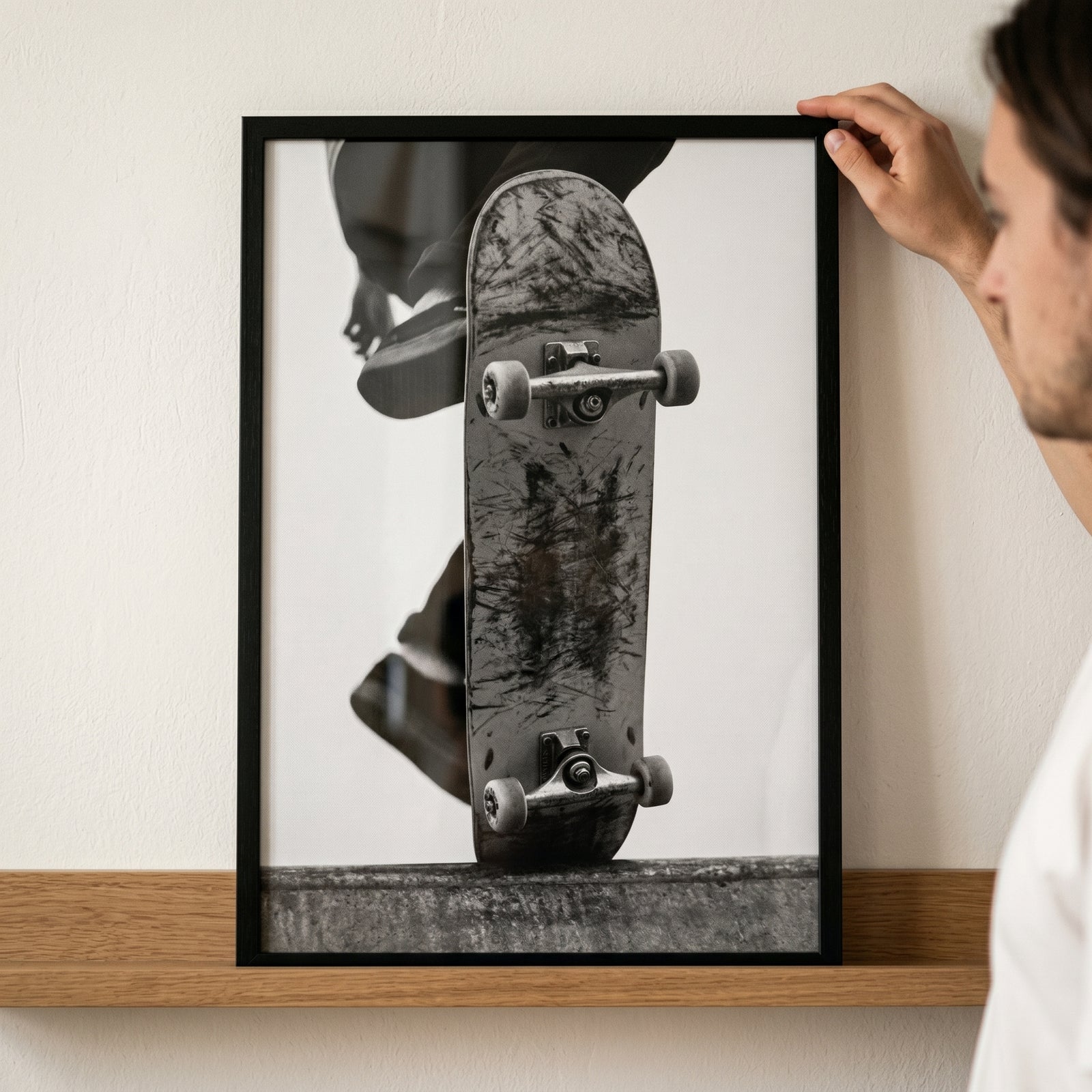 Framed scratched deck vintage skateboard artwork held by a hand against a white wall over a wooden shelf.