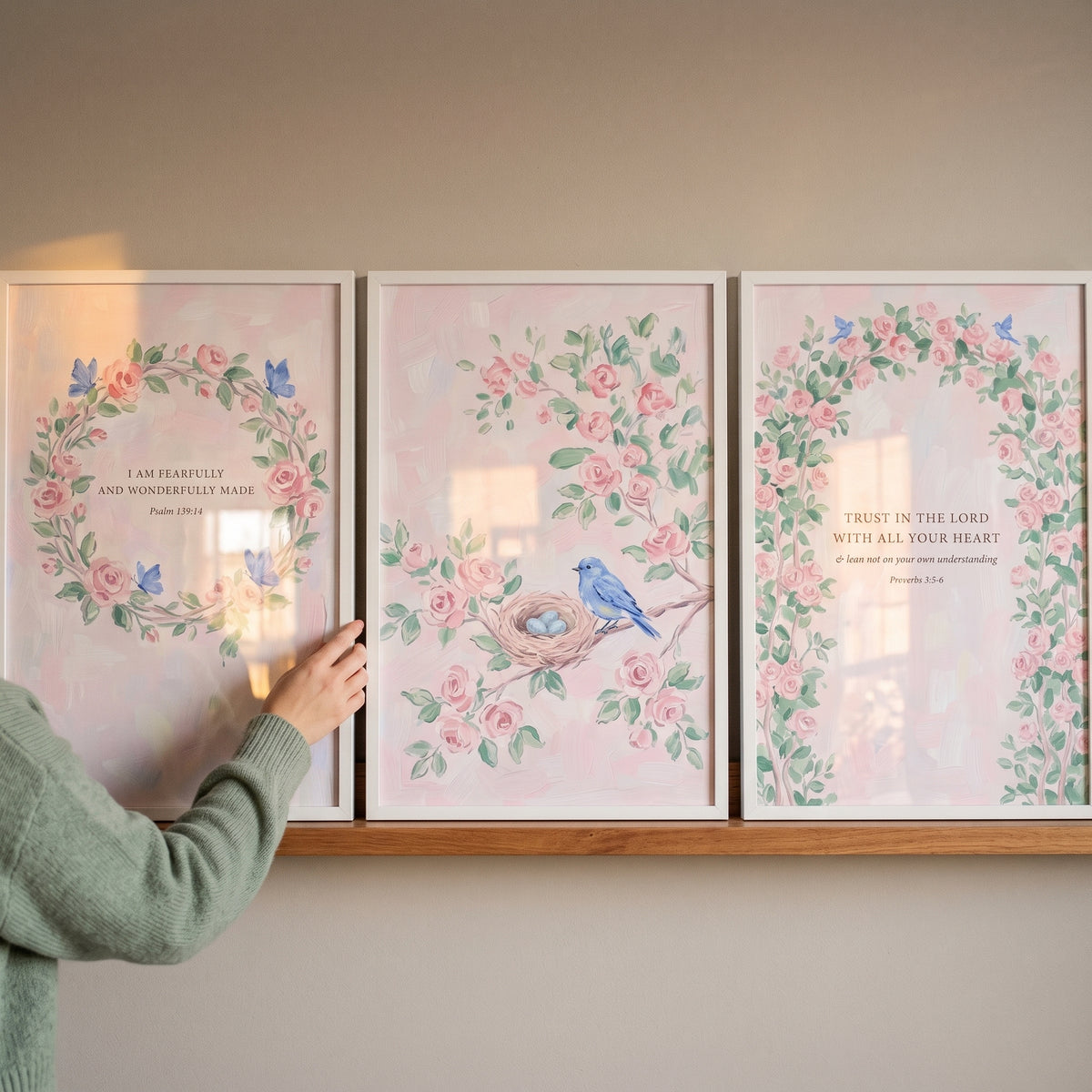 Hand arranges a scripture blush pink rose wall art set of 3 featuring birds and butterflies on a wooden shelf.