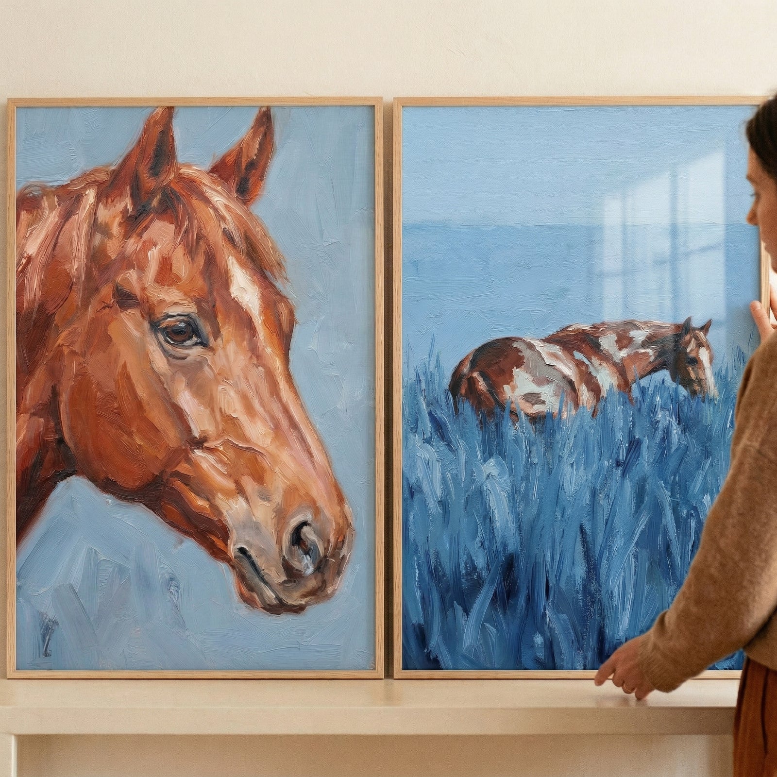 Two framed textured horse paintings displayed side by side, one showing a close up portrait of a brown horse against a soft blue background and the other showing a horse resting in tall blue grass. A person adjusts the artwork, suggesting styling in a calm rustic home setting.