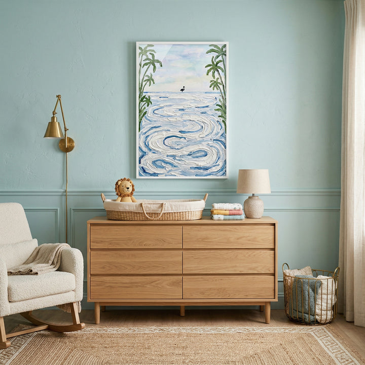 Framed painting of swirling ocean waves with a small bird near the horizon hangs above a wooden dresser in a soft blue nursery. A changing basket lamp rocking chair and woven textures create a calm coastal inspired space centered around the artwork.